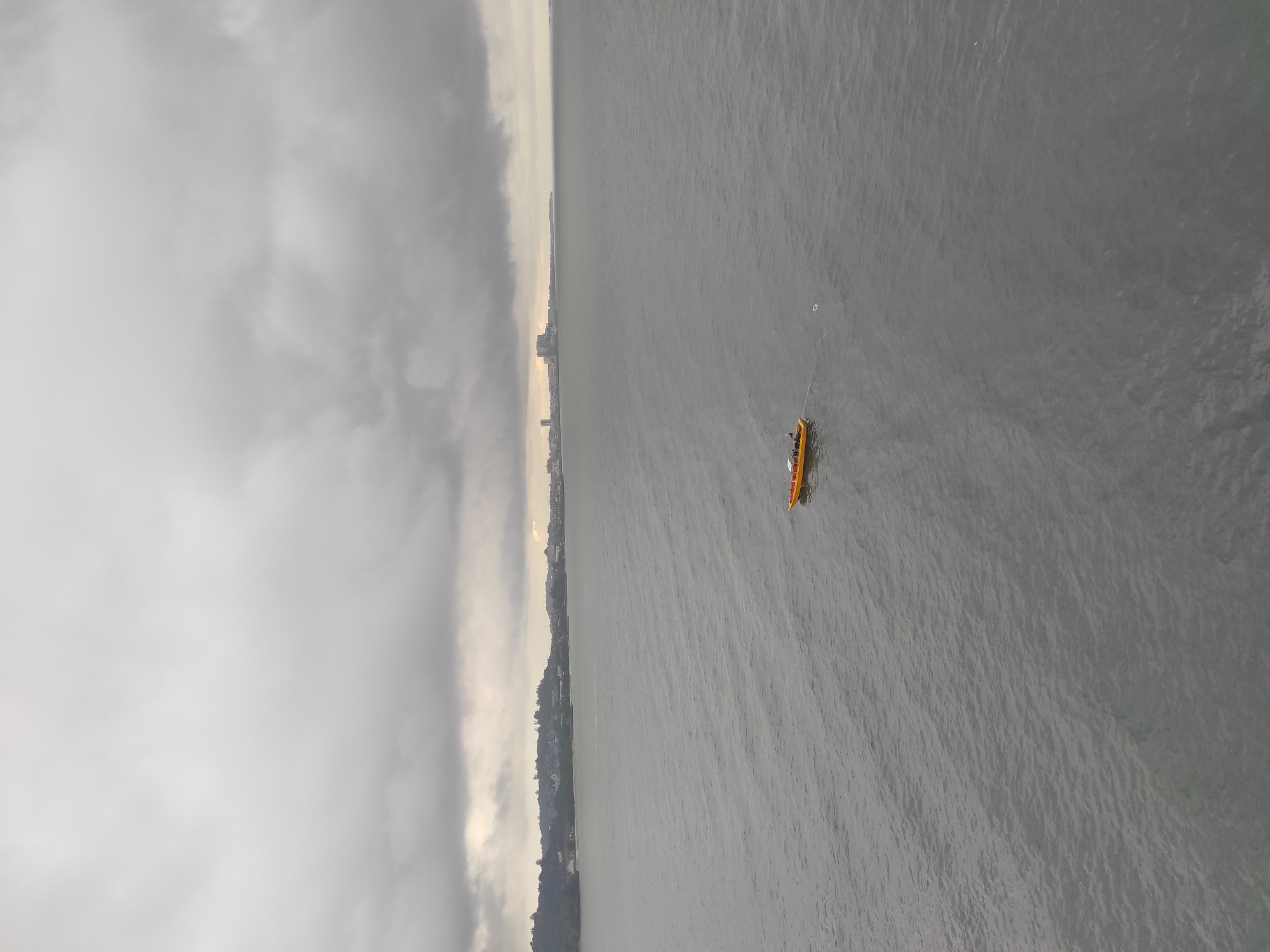 An overhead, wide shot captures a calm, choppy, gray body of water under an overcast sky. In the middle of the frame, a small, yellow boat with an orange trim sails across the water, tethered to a long rope that trails behind it. The boat contains at least two visible figures. In the background, a distant shoreline with a scattering of buildings and trees is visible, extending across the horizon. The overall scene is one of a muted, gray day on the water.