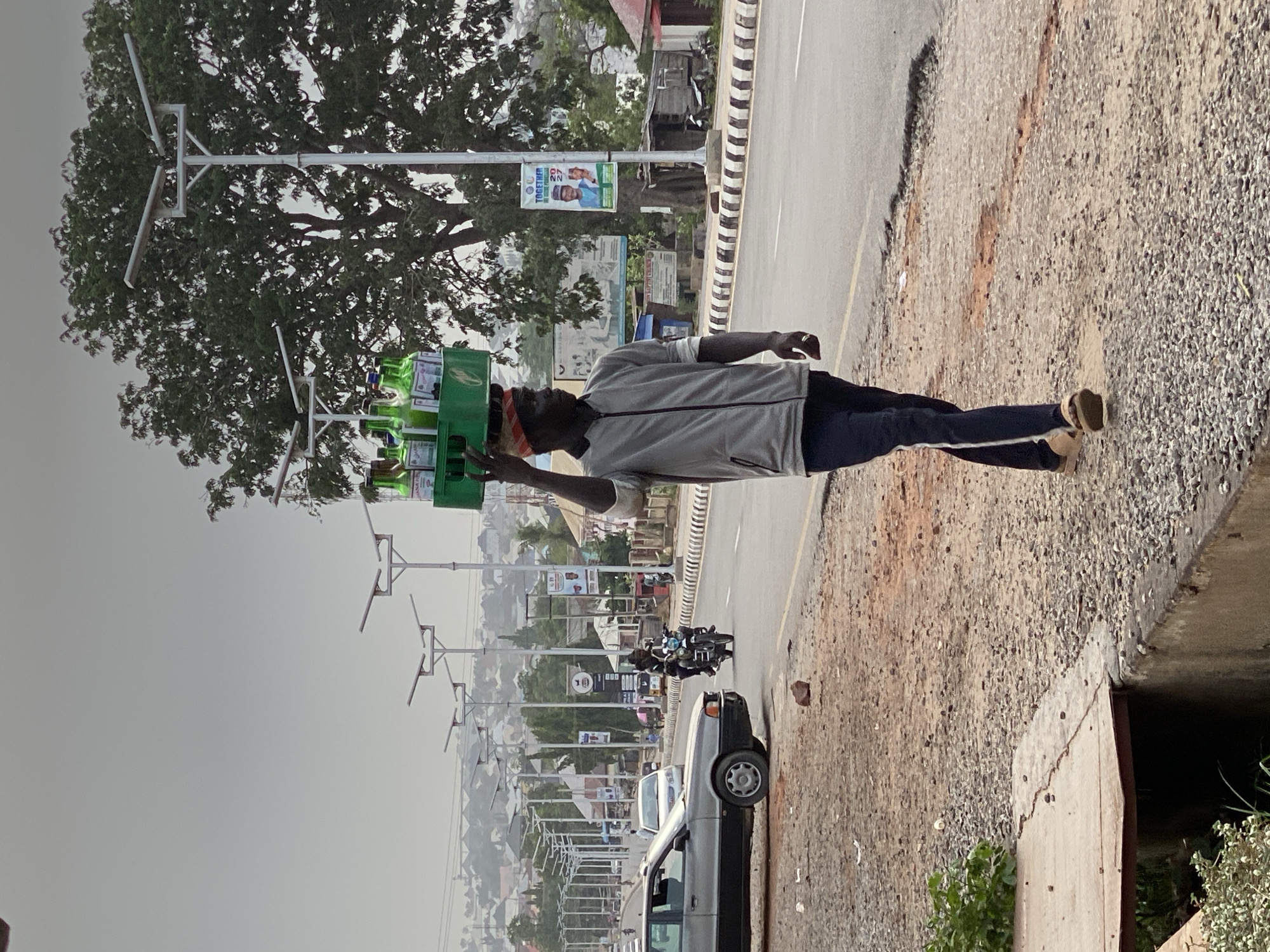 This image captures a street scene in Adankolo, Nigeria, under what appears to be a hazy or overcast daytime sky. The primary subject is a man walking along the gravel shoulder of a paved road, skillfully balancing a green crate filled with bottles, likely beverages, on his head. He is dressed in a light grey long-sleeved shirt, dark trousers with a white stripe down the side, and light-colored sandals. A red and white patterned band is visible on his head beneath the carried crate.
To the man's right, a paved road is bordered by a black and white striped curb. Further down the road, a silver car is parked, and a motorcycle carrying multiple individuals is in motion. The background features a series of modern streetlights, each equipped with solar panels, lining the road. Large, dense trees and distant buildings are also visible, suggesting an urban or semi-urban environment. The immediate foreground shows a rough, gravelly terrain, indicating the edge of the road or a bridge structure.
A notable detail is a political campaign poster affixed to one of the street poles. The poster displays images of two men, presumably candidates, alongside text that includes "TOGETHER WE CAN DO MORE" and the number "27". Partially visible names include "SENATOR AHMED..." and "Dr. YINUSA...". The scene depicts everyday activity with elements of local infrastructure and political campaigning.