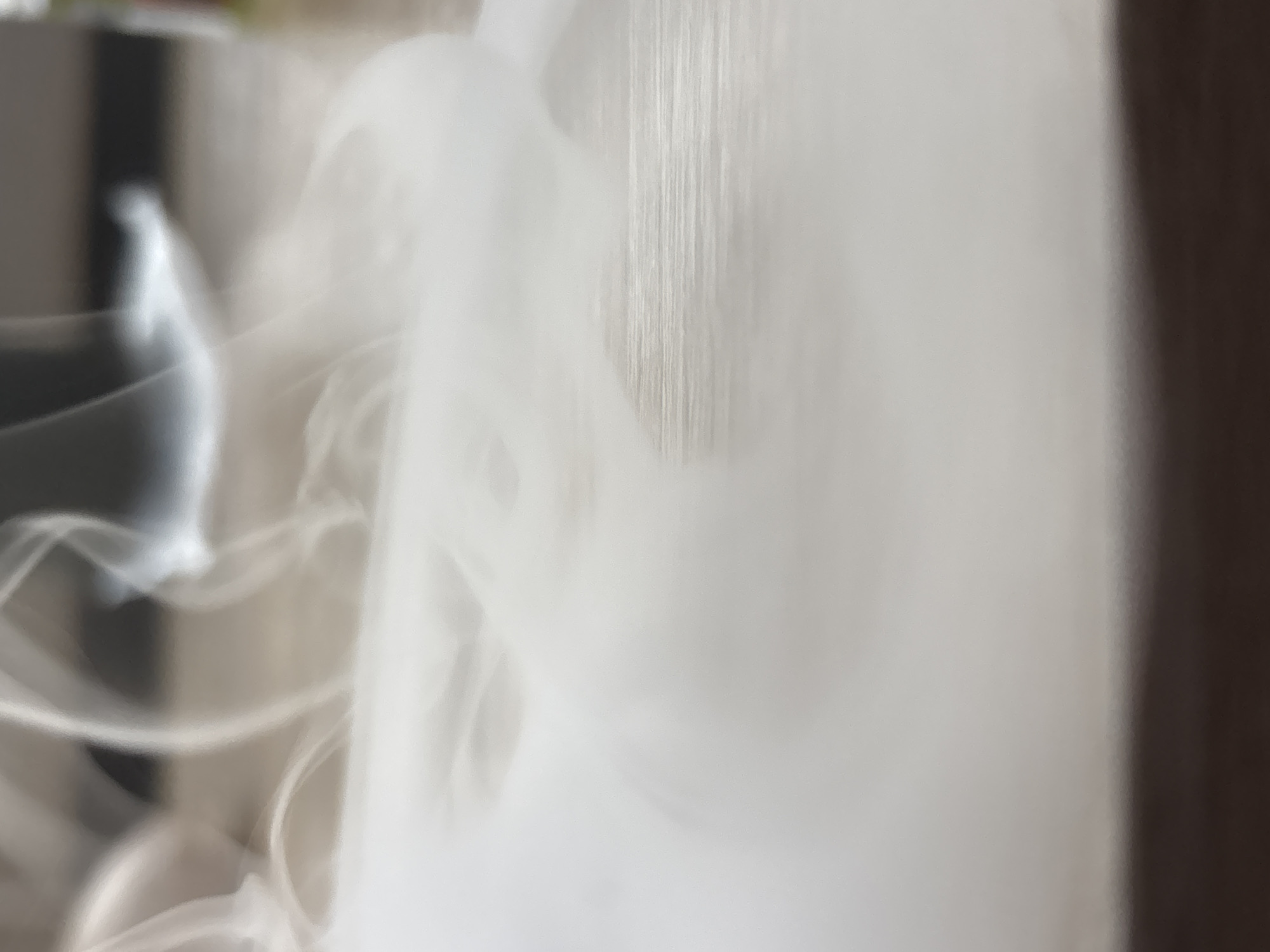 The image depicts a close-up, abstract view of white smoke or vapor swirling over a textured surface, possibly wood grain or a similar material. The lighting is soft and diffuse, creating a hazy and ethereal atmosphere. The top portion of the image is blurred and shows indistinct shapes and colors, suggesting a background that is out of focus. There are no discernible landmarks or specific environmental cues that would place this scene in Saint Petersburg, Russia, or any other specific city. The image is primarily focused on the texture and movement of the smoke.