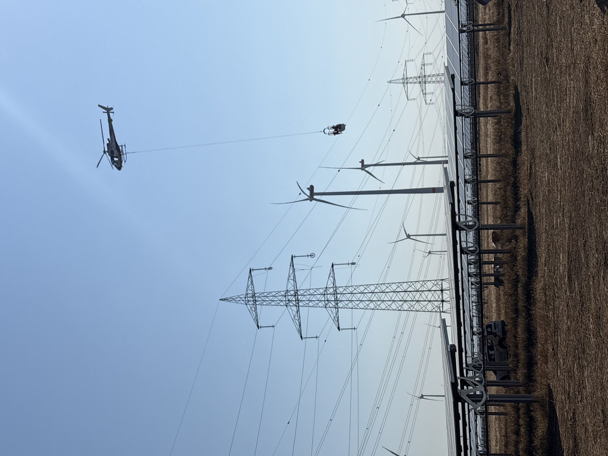 This image depicts an aerial view of a solar farm and wind farm in Neusiedl am See, Austria. A helicopter is flying in the sky, carrying a small platform with two individuals suspended from it by a cable. The platform appears to be moving along a power line, possibly for inspection or maintenance purposes. In the foreground, rows of solar panels are arranged on metal racks, with a utility vehicle parked among them. Several wind turbines are visible in the background, along with high-voltage power lines and transmission towers. The sky is clear and blue, suggesting fair weather. The ground is covered with dry grass and soil. The scene captures a moment of industrial activity involving renewable energy infrastructure.