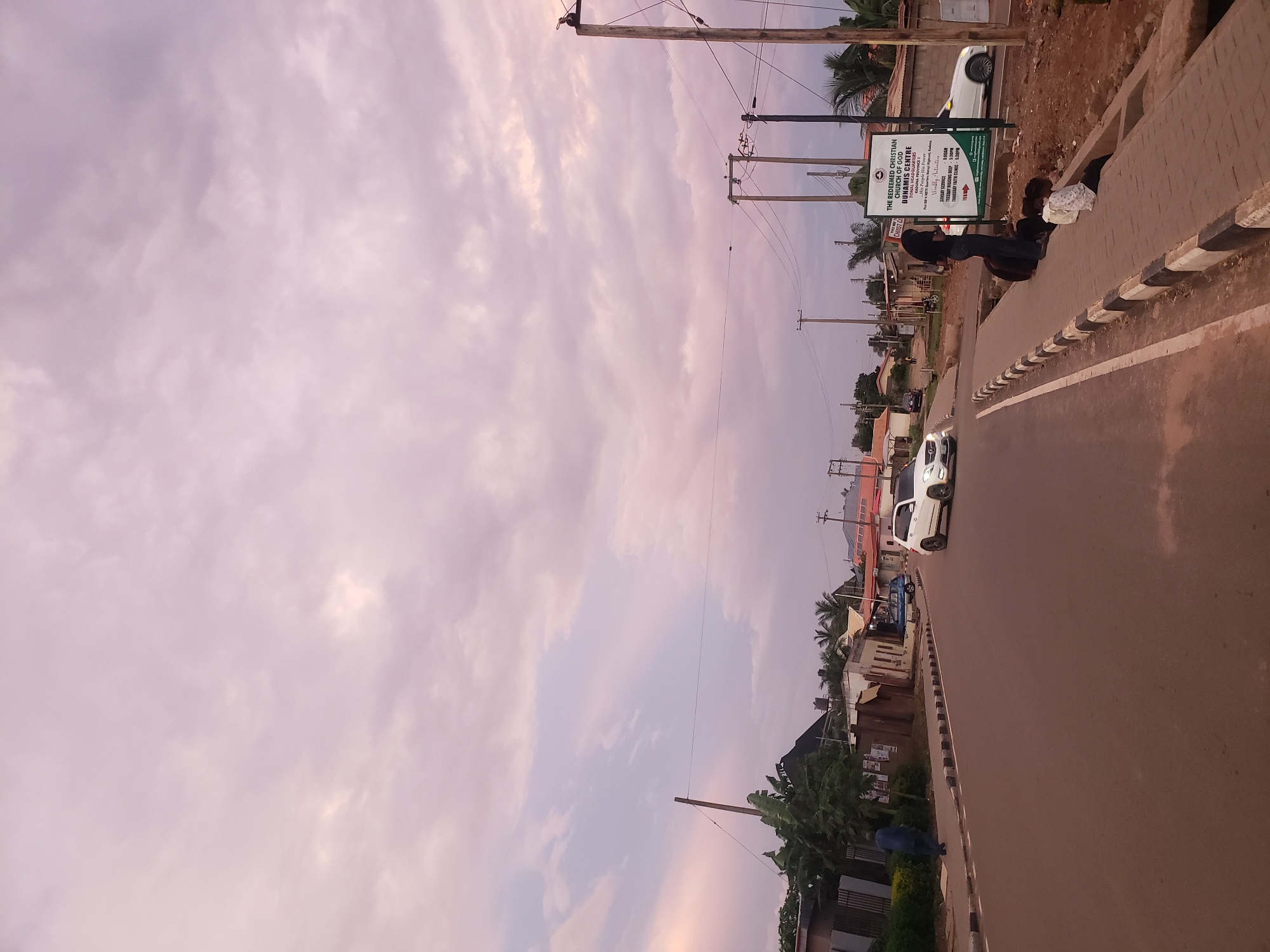 The image depicts a street scene in Narayi, Nigeria. The sky is filled with soft, pinkish-purple clouds, suggesting either dawn or dusk. The street itself is paved with asphalt and has a sidewalk with black and white striped curbing.
On the left side of the street, there are buildings with corrugated metal roofs, some with visible advertisements posted on their walls. Vegetation, including banana trees, grows along the sidewalk. A dark-clothed person is walking away from the camera on the sidewalk. A blue car is parked in front of a building.
On the right side of the street, there are several utility poles with electrical wires. A large sign is prominently displayed, which reads "THE REDEEMED CHRISTIAN CHURCH OF GOD DUNAMIS CENTRE, KADUNA PROVINCE 2". Below this, service times and other information are listed. Several people are seated or standing on the sidewalk near the sign. One individual is wearing a black top and jeans, and two younger individuals are sitting on the edge of the sidewalk. A white SUV is driving away from the camera on the road, and another white car is parked further down the street. The environment appears to be a mix of residential and commercial structures.