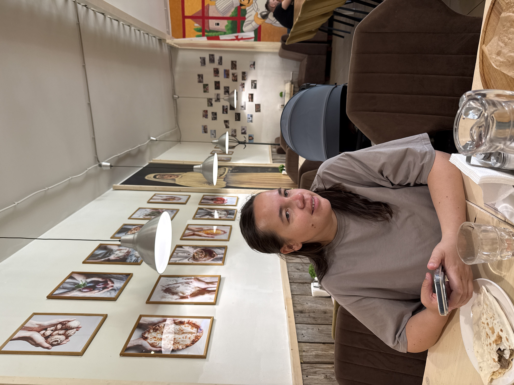 A medium-shot view captures a woman seated at a table inside a restaurant. The woman, centered right, is wearing a taupe shirt and holding a smartphone in her right hand. On the table, beside her, is a plate with a flatbread, a glass, and some other tableware.
In the background, which provides depth and context to the scene, several framed pictures hang on the wall, displaying various food-related images. Above the pictures, a long painting stretches across the wall, adding an artistic element to the restaurant's decor.
The restaurant is dimly lit, with pendant lamps hanging down to provide illumination. Additional furnishings, including brown upholstered chairs and a stroller, are visible, suggesting a family-friendly environment.
While the city of Moscow, Russia, is not explicitly confirmed by visual elements alone, the style of decoration and the ambiance of the restaurant could be consistent with dining establishments in a major European city.