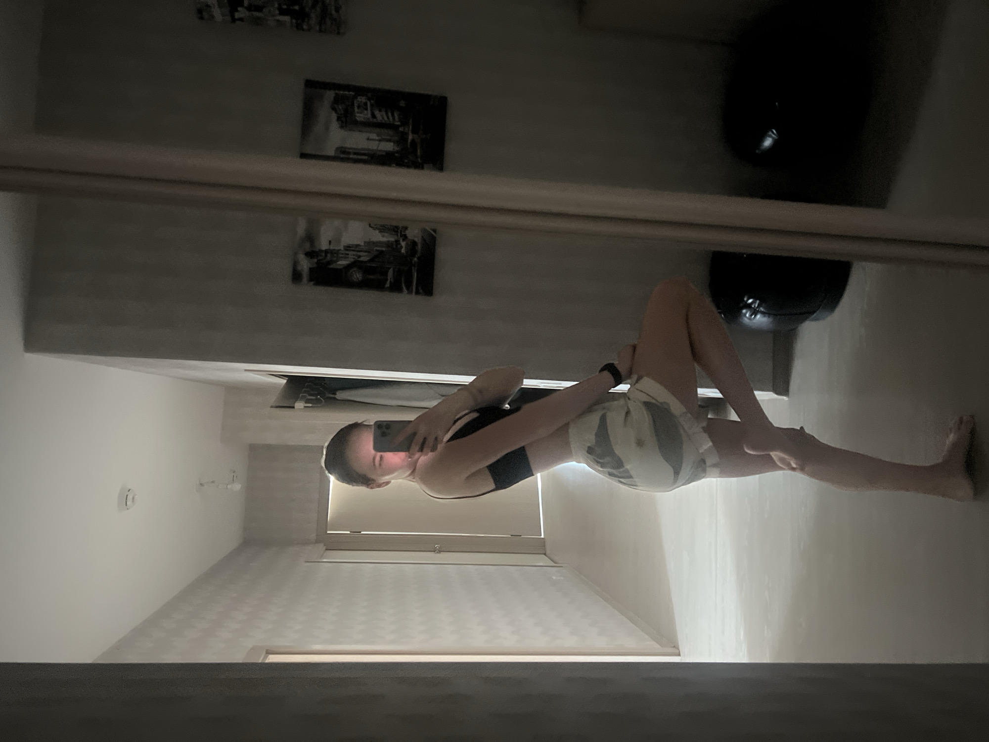 A fair-skinned person with short hair takes a picture with a mobile phone in front of a mirror. The person is wearing a black sports bra and patterned shorts. The person is lying on their side with one leg extended. The reflection shows a hallway with a light fixture and a fire alarm. There are framed black and white pictures on the wall and two black exercise balls in the background. The location is a private residence, the city of Perm, Russia cannot be ascertained.