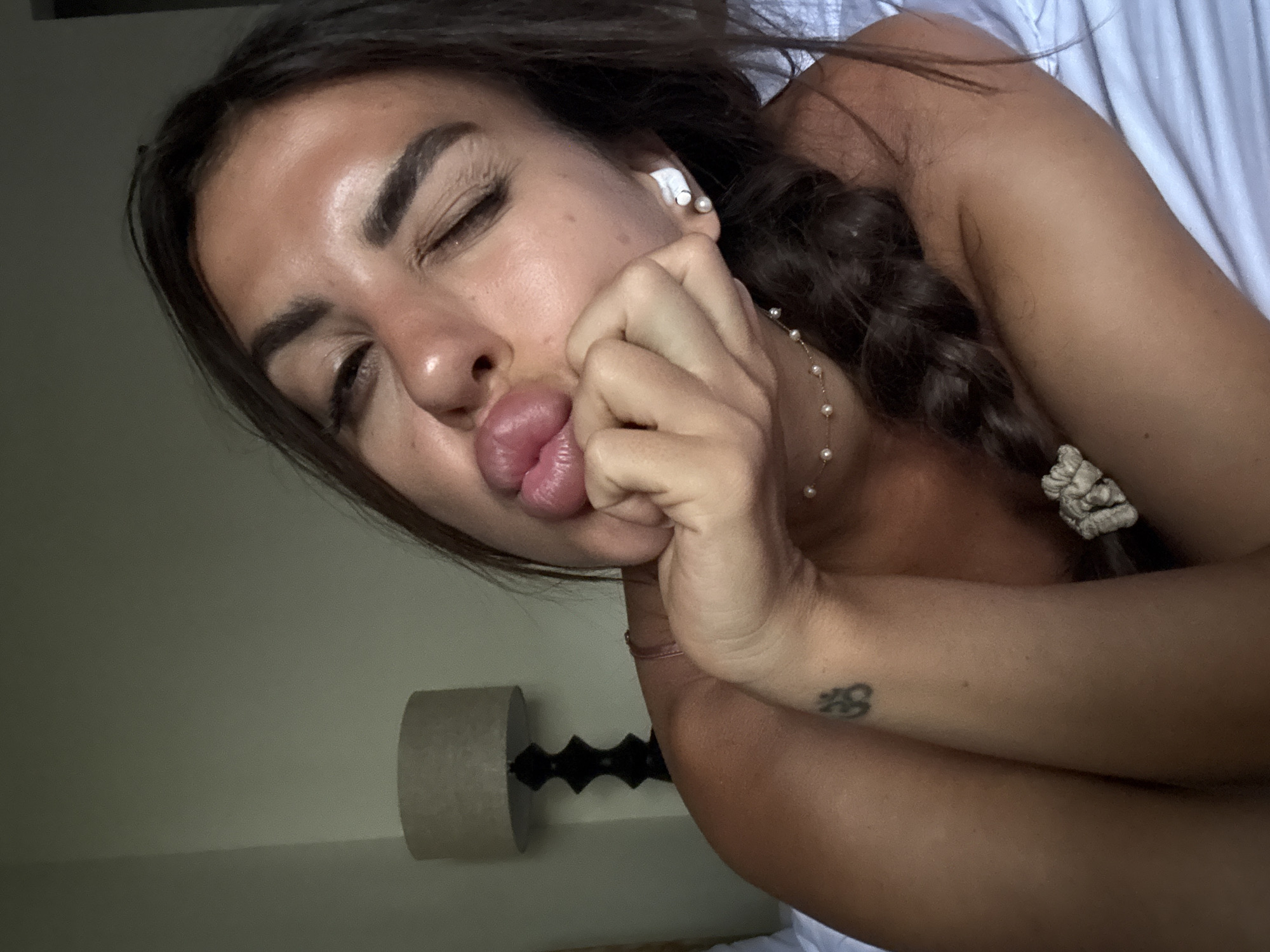 A woman is lying on a white sheet in North Kuta, Indonesia, with her eyes closed and lips puckered. She is wearing an earbud in her left ear, and a necklace of pearls. Her brown hair is braided, held with a white scrunchie. There is a tattoo on her forearm. A cylindrical lamp stands nearby.