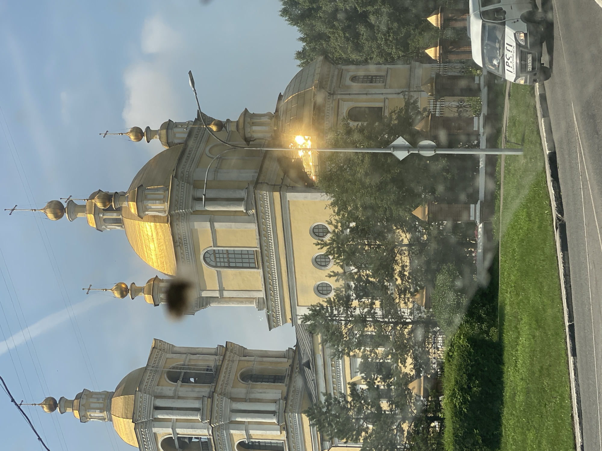The image captures a sunlit daytime view of the Novokuznetsk City Central Mosque in Novokuznetsk, Russia. The mosque features a prominent yellow facade and multiple golden domes, one of which is brightly reflecting the sunlight. Adjacent to the mosque is a bell tower. In the foreground, a white van with Cyrillic lettering on its windshield is driving on a paved road. The scene is framed by green grass, manicured hedges, and a decorative fence with golden-topped pillars. Trees line the background behind the mosque. The image appears to be taken from a moving vehicle, as evidenced by the slight blur and the presence of what look like water droplets or smudges on the surface from which the photo was taken.