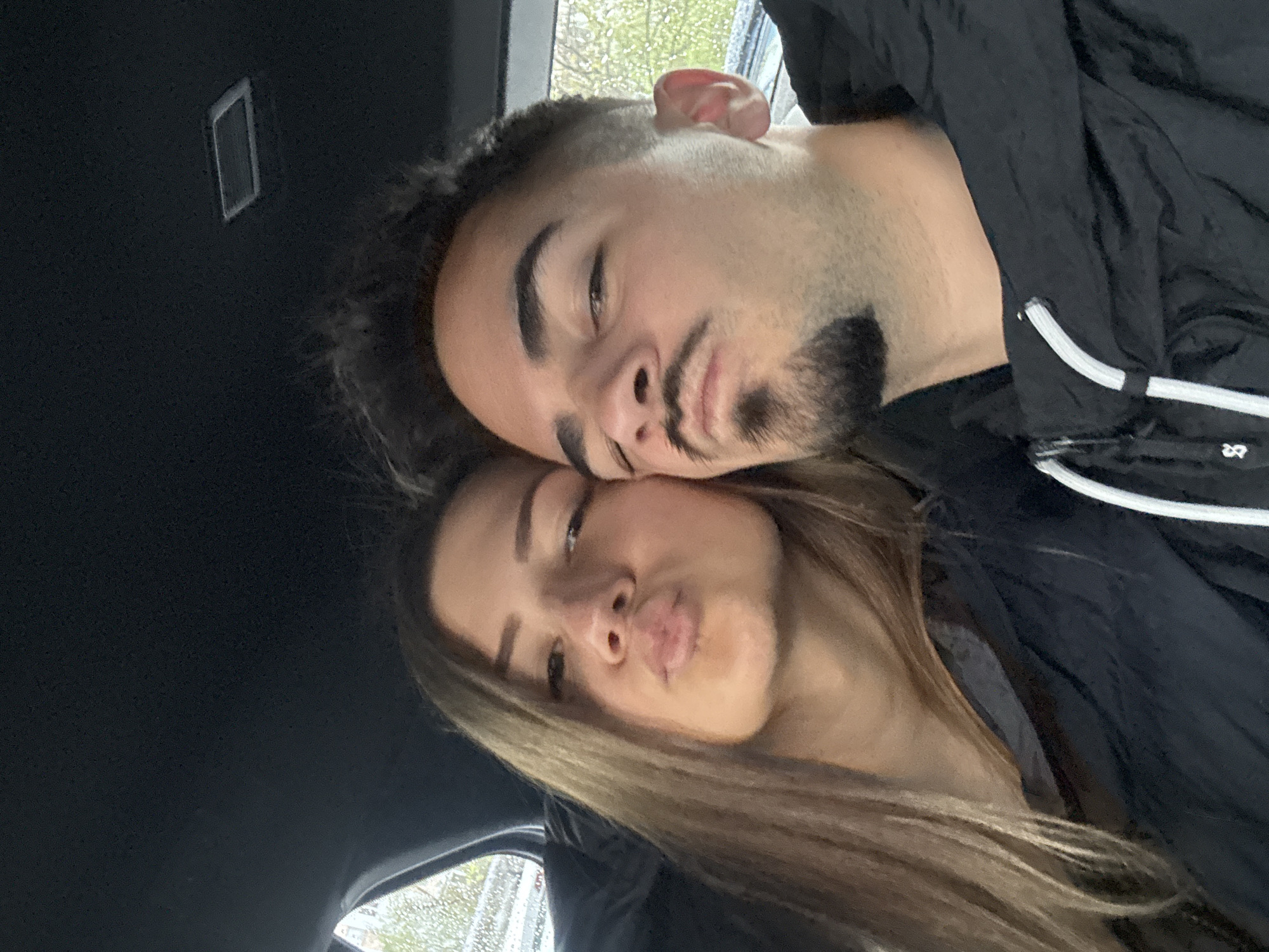 A photo, taken from a low angle, depicts two people. A woman with long brown hair is in the foreground, making a kissing face towards the camera. Behind her, a man with short dark hair and a beard rests his head against hers. They are in what appears to be a car, based on the dark ceiling and side window visible, with rain droplets visible on the glass. The man is wearing a dark jacket with white drawstring accents. The specific location in Sofia, Bulgaria is impossible to determine from the visual information.
