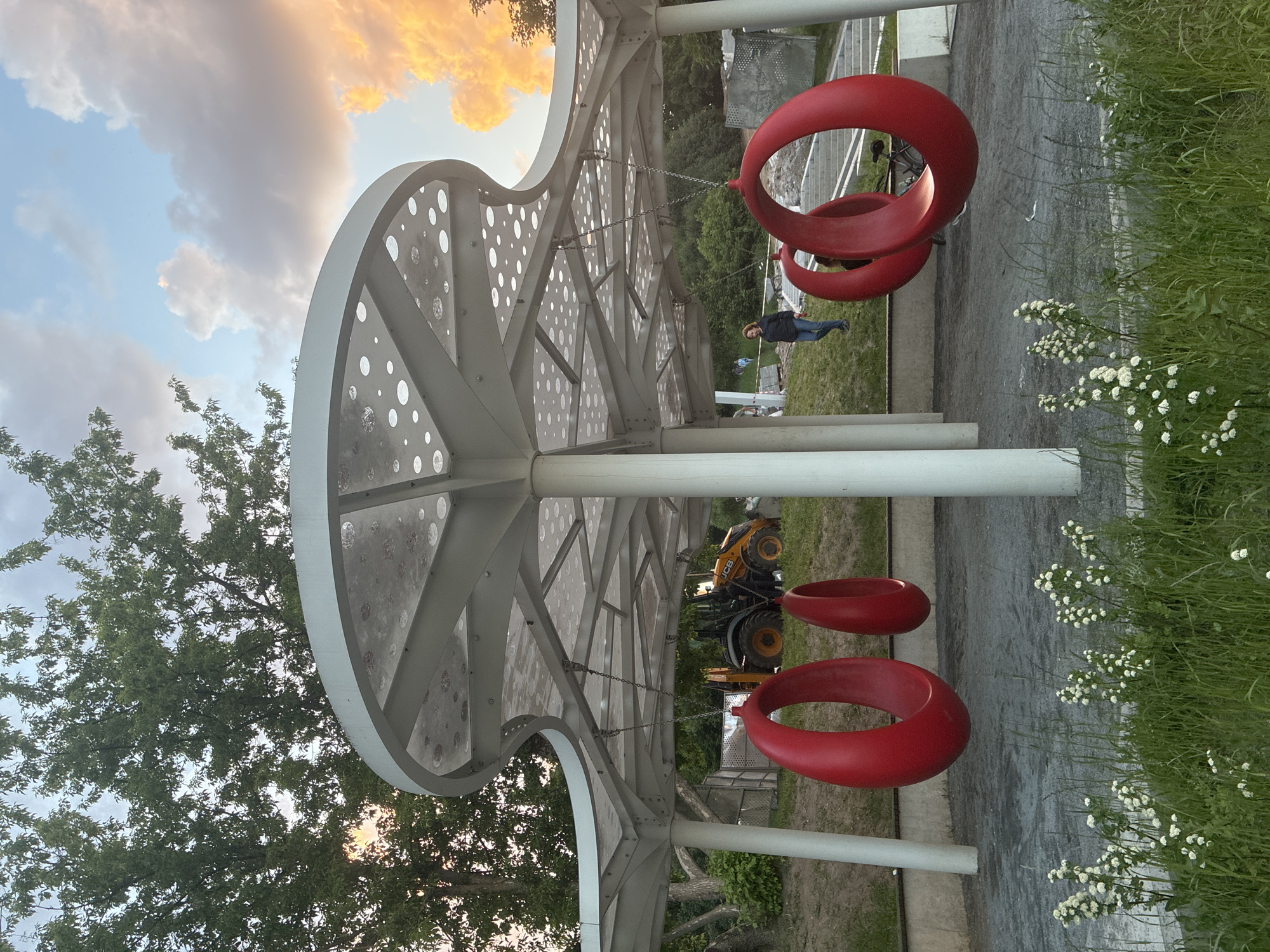 This image depicts a vibrant outdoor public space, likely a park or recreational area, in Moscow, Russia, during the golden hour of either sunrise or sunset. The sky displays a mix of soft blue and scattered clouds, with some illuminated by a warm, orange glow.
The main subject is a contemporary, white architectural canopy or pavilion with a distinct arched design. Its surface is adorned with various-sized circular perforations, and its underside reveals a complex, geometric truss structure made of white metal.
Beneath this canopy, several unique, bright red, donut-shaped swings are suspended by chains. One person is visible standing to the right of the swings, dressed in dark attire, seemingly observing the surroundings. The ground features a combination of grey paved areas and green grass. To the right, tall green grass with small white flowers adds a touch of natural beauty.
In the background, numerous green trees frame the scene. A yellow JCB backhoe loader is partially visible further back, hinting at nearby construction or maintenance activities. The scene conveys a tranquil yet modern atmosphere, blending architectural design with natural elements. No visible text is present in the image.