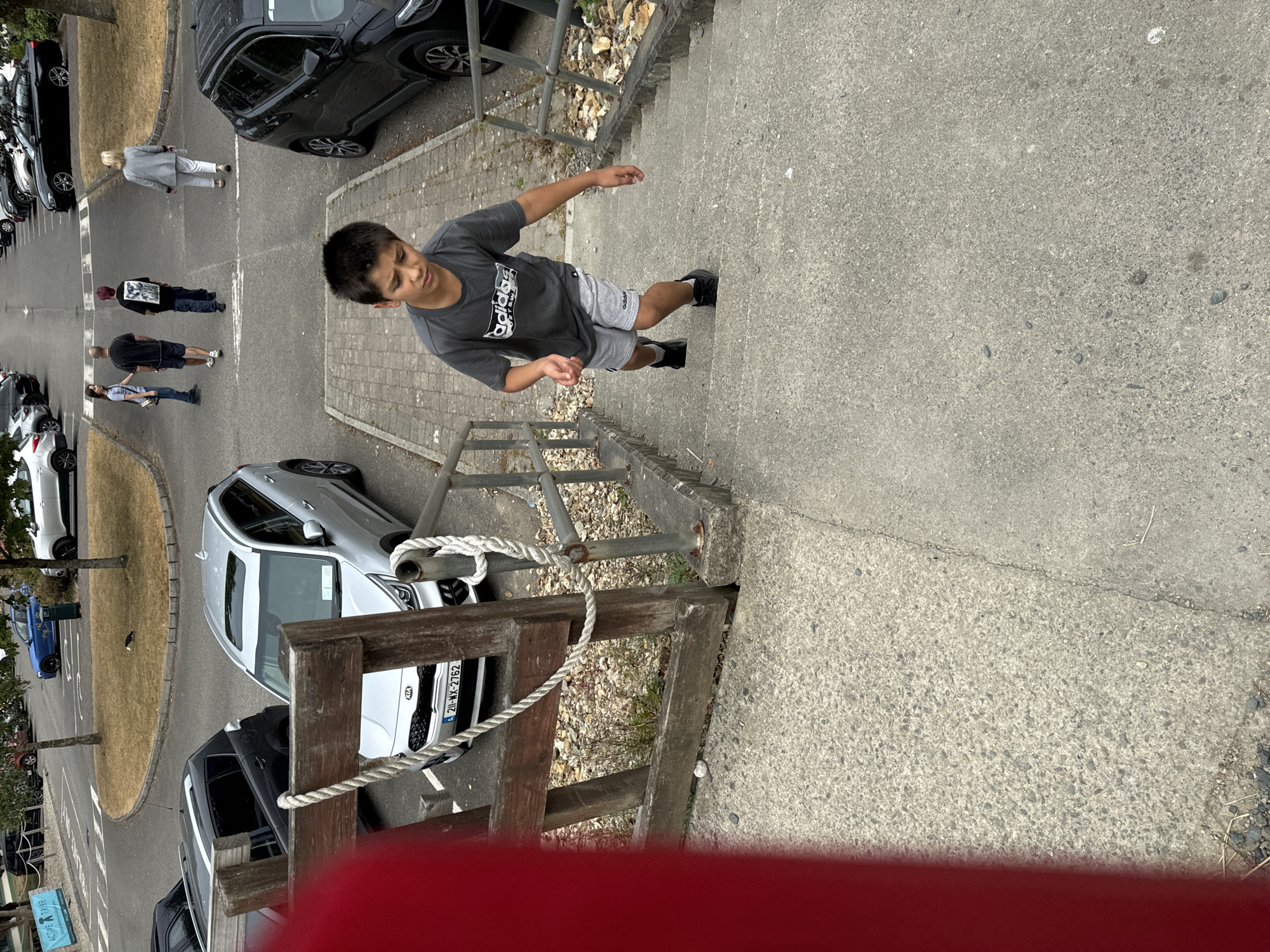 A young boy is climbing a concrete staircase outdoors. He is wearing a grey t-shirt with an Adidas logo and grey shorts, and black sneakers. The boy is looking down as he walks up the stairs. In the background, several cars are parked in a lot, and some adults are walking in the distance. The environment appears to be a park or recreational area in Courtown, Ireland, with trees and grass visible.