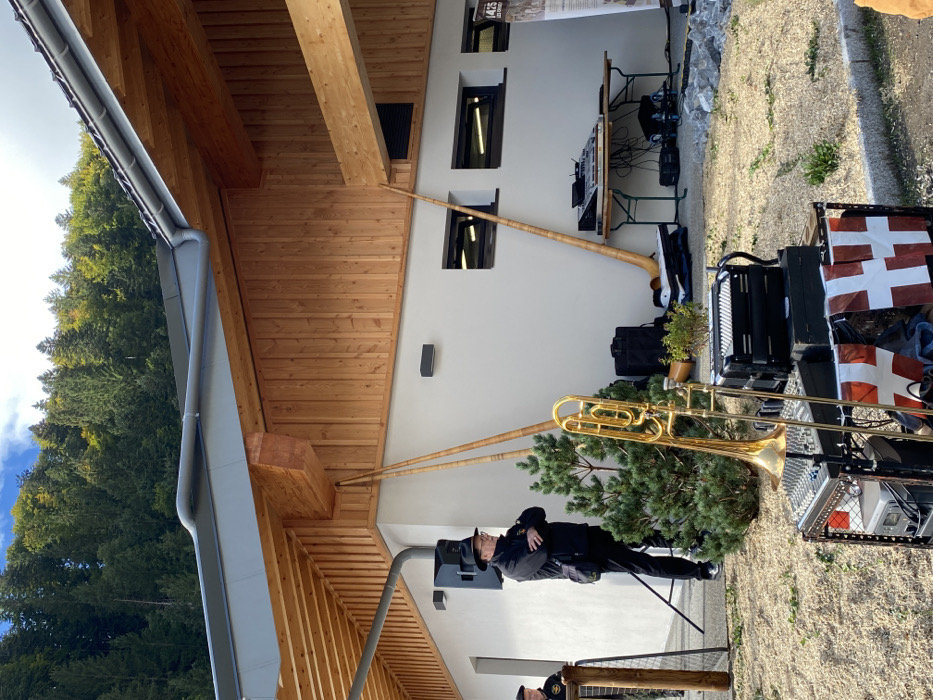 This image depicts an outdoor event setup against a modern building in Abondance, France. The building features a grey tiled roof with a prominent gutter, an overhanging eave with a visible wooden underside, and a wall partially clad with vertical light-brown wooden planks and partially with plain white stucco. Two rectangular dark-framed windows are visible in the white stucco section of the wall, along with two black wall-mounted light fixtures.
In the background, a dense green forest covers a steep hillside under a blue sky with scattered clouds. The ground in the foreground and mid-ground is covered with light-colored gravel.
Musical equipment is prominent. Three long, light-brown wooden alphorns lean against the building: two on the left side, angled towards the wooden cladding, and one on the right side, leaning against the white stucco. In the foreground, slightly left of center, a golden brass trombone stands on a metallic rack holding a sound mixer. Below the trombone, a black accordion rests on a box. A banner, consisting of several small white flags with red crosses, is draped across the front of this equipment stand. Further right, a collapsible wooden table supports a larger mixing console and other audio equipment, with cables visible on the ground. A black speaker is positioned on the ground to the right of this table. Another black speaker is mounted on a stand to the left of the primary human subject.
One main male subject is visible in the mid-ground, slightly left of center. He is wearing a black hat, glasses, a black jacket, and dark trousers, with a dark shoulder bag. He stands with his arms crossed, looking towards the right. Part of a second male subject is visible on the far left, wearing a dark beret and a dark jacket with a gold emblem, standing behind a wooden post.
A small, conical evergreen bush is situated between the main male subject and the foreground musical equipment. On the far right, a narrow vertical banner with "1425 LES CROSES" is affixed to the building's wall.