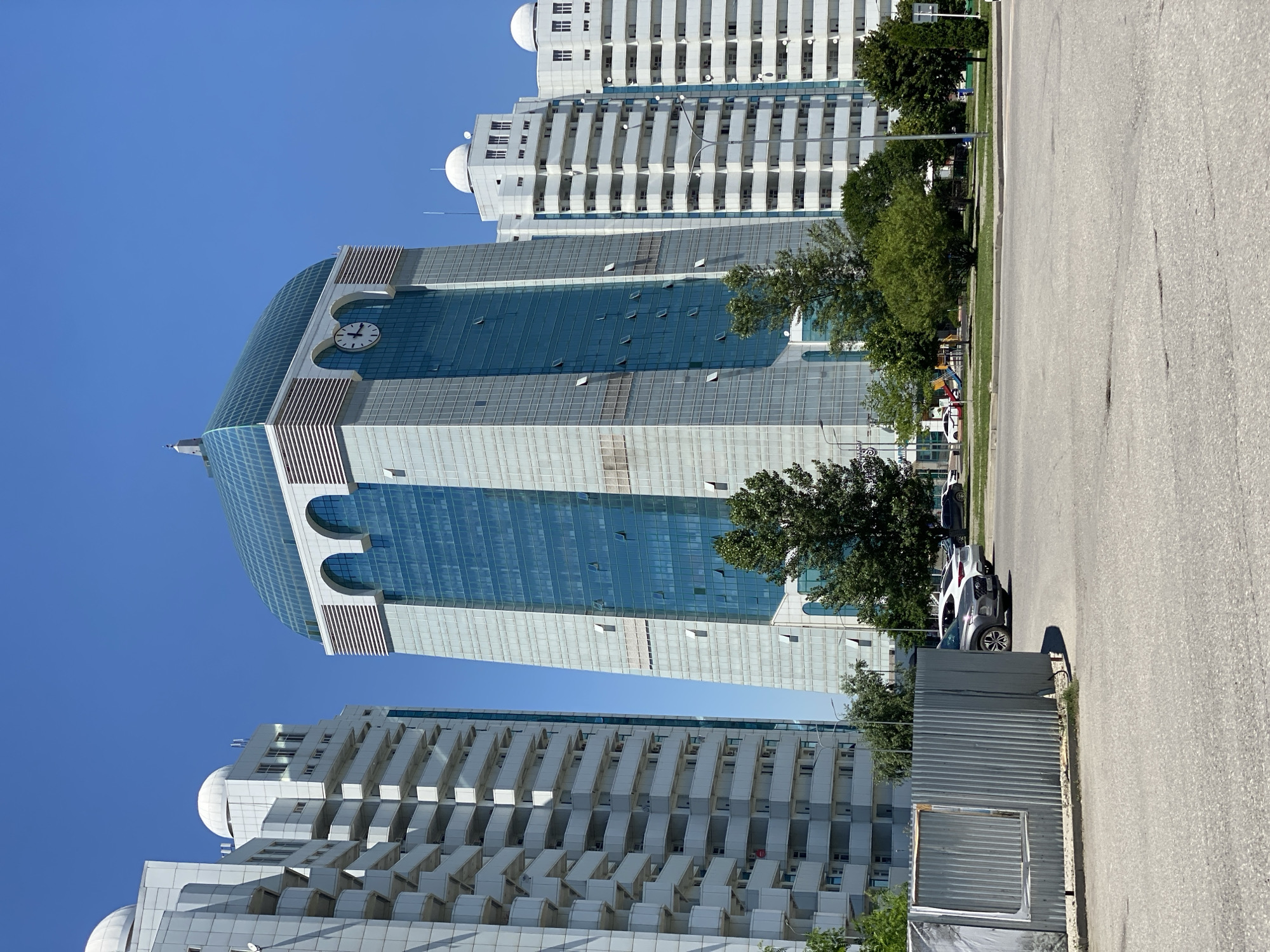 This is a vertical, eye-level photograph taken outdoors on a sunny day, likely in the afternoon due to the clear blue sky and strong shadows. The scene features a modern cityscape dominated by tall, white apartment buildings and a prominent glass-fronted office building with a large clock face near its top. The apartment buildings have many balconies, giving them a layered appearance. The office building has a curved glass facade in shades of blue and grey, with distinct architectural features like arched window openings.
In the foreground, a paved road runs vertically along the right side of the image, with a sidewalk or grassy area separating it from the buildings. A white car is parked on the side of the road, casting a shadow. Some of the windows on the office building are open. To the right of the office building, mature green trees provide a natural element. A temporary metal fence is visible in the middle ground.
There are no people visible in the photograph, and the overall mood is one of urban development and quiet activity. The location is indicated as Shali, Russia, which provides a geographical context for the modern architecture. The time of day is suggested by the bright sunlight and sharp shadows. The weather appears to be clear and warm. There is no visible text in the image.
