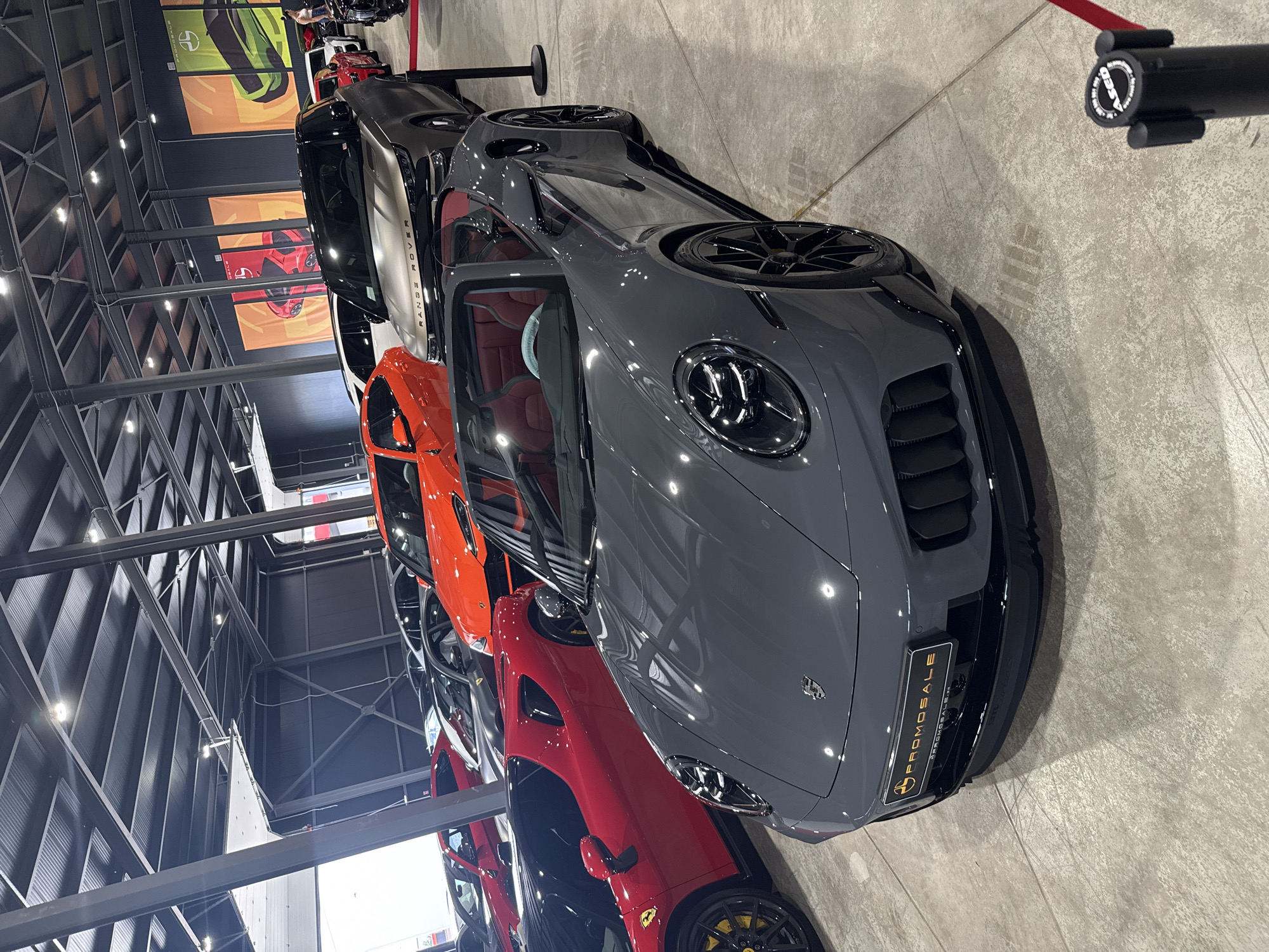 The image shows an indoor exhibition space, likely a car showroom, with multiple luxury vehicles on display. The foreground is dominated by a grey convertible Porsche 911, with its sleek design and red interior clearly visible. To the left, a vibrant red Ferrari is parked. Further back, an orange Lamborghini SUV and a grey Range Rover are among other cars. Large banners with car graphics hang on the wall in the background. The ceiling features industrial metal beams and lighting fixtures. The floor is a polished concrete surface. The location is identified as a car exhibition in Plovdiv, Bulgaria, specifically within a modern, spacious venue. There are no people clearly visible interacting with the vehicles, suggesting a static display. The overall impression is of a high-end automotive event.