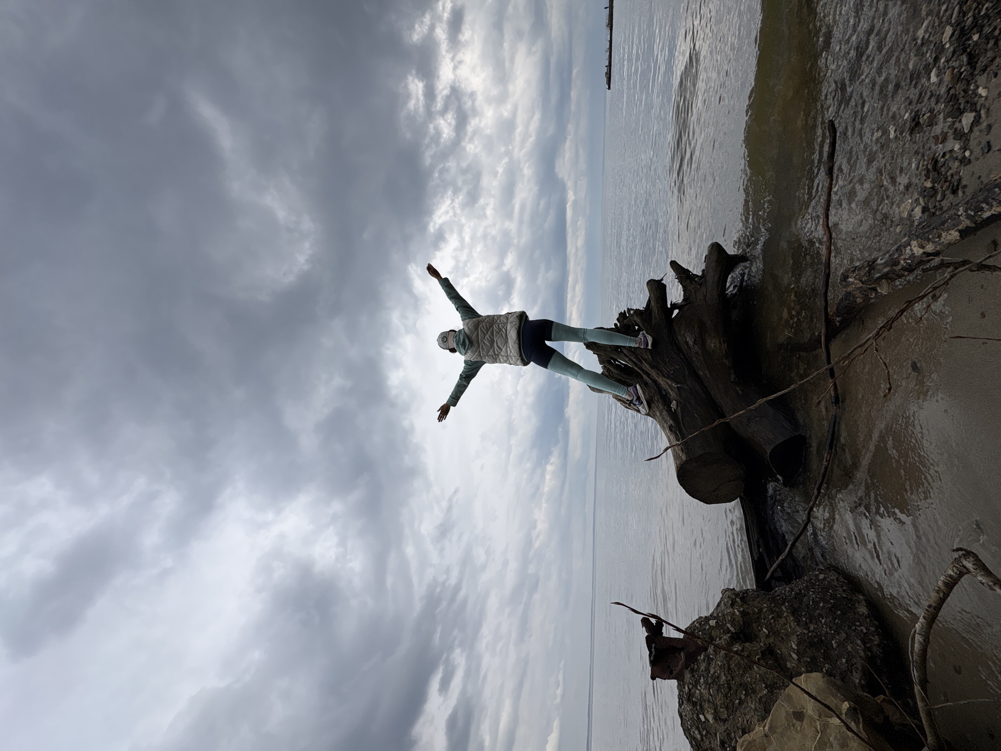 A woman is standing on a tree stump on a beach with her arms outstretched. The sky is cloudy and the water is visible in the background. The location is likely the city of Ulyanovsk, Russia.