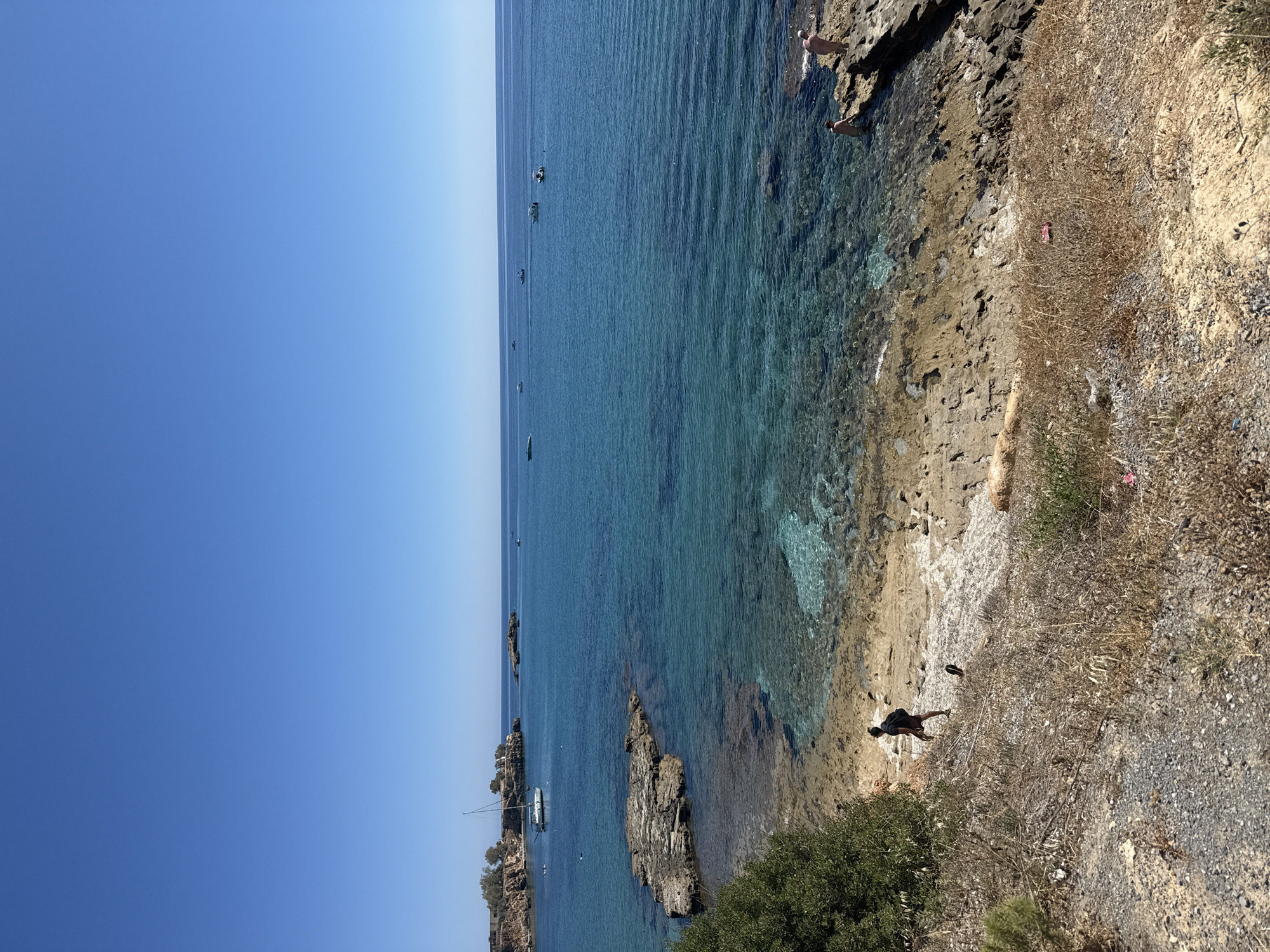 The image depicts a coastal scene in Chersonisos Municipal Unit, Greece, on a clear, sunny day. The vast, blue sky meets a calm, turquoise sea, with the water's clarity allowing visibility of the rocky seabed near the shore. The coastline is rugged and dry, with sparse vegetation and rocky outcrops.
Several small boats are visible in the distance on the sea, suggesting a peaceful day for boating or swimming. Near the shore, there are a few people. One person is standing on the rocky shore, another is in the water near the rocks, and two individuals are further out in the sea. On the dry, rocky land, there are two people. One is standing near the water's edge, and another is further inland on the dry, scrubby terrain. There's a flip-flop visible on the ground.
The overall mood is tranquil and summery, conveying a sense of relaxation and enjoyment of a seaside environment. The lighting suggests it is daytime, likely late morning or afternoon, given the bright sunlight and clear conditions. The location cues of the rocky coastline, the clear blue water, and the presence of boats are consistent with a Mediterranean coastal setting. No text is visible in the image.
