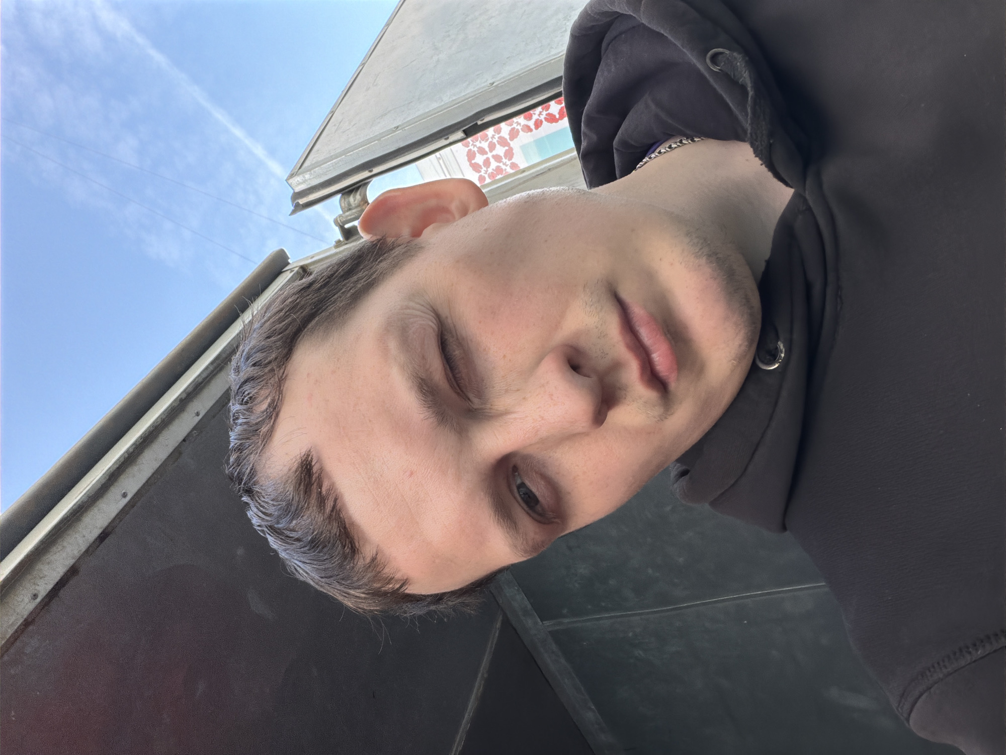 A man wearing a black hoodie is positioned in front of a vehicle, possibly a truck, in what is potentially Chaikovsky, Russia. The sky, visible in the background, is light blue. The man has his left eye squinted or closed, appearing to wink. He has short brown hair and some stubble. A silver chain is visible around his neck. The vehicle behind him appears to be grey and metallic. A window with what may be floral decorations is slightly visible above the man's head.