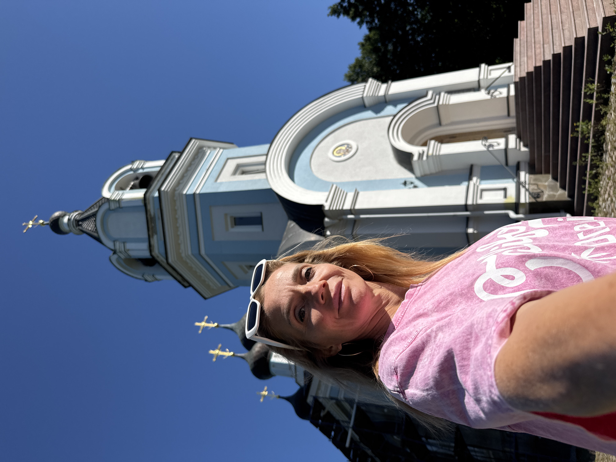 A woman is taking a selfie with a church in the background in St. Ozerki, Russia. She is wearing a pink t-shirt and white sunglasses pushed up on her head. The church has blue and white accents and a bell tower with a gold cross on top. The sky is clear and blue.