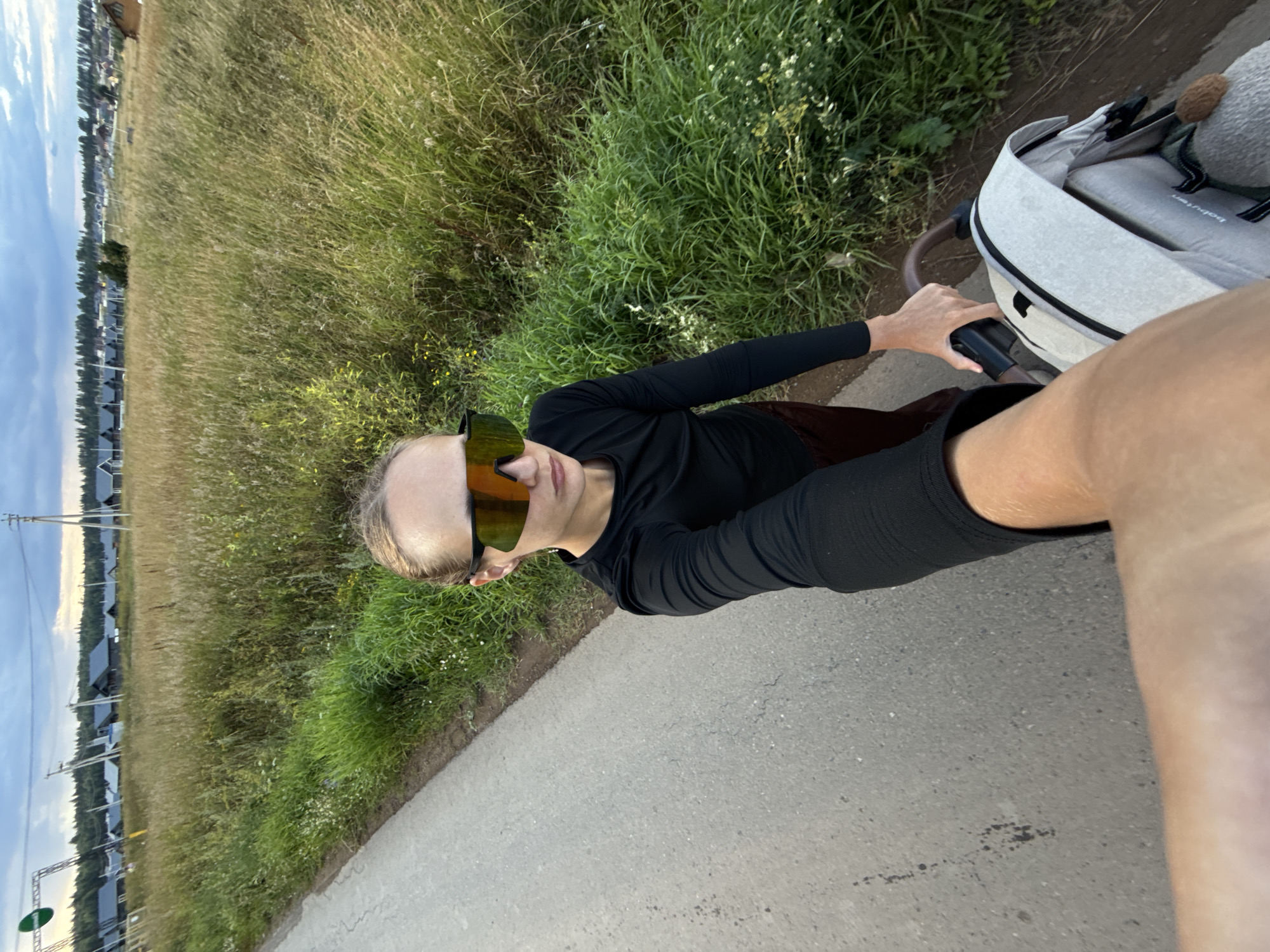 A person is pushing a stroller on a paved path next to a grassy field. The person is wearing a black long-sleeved shirt and sunglasses with orange lenses. The stroller is light gray with a brown handle. In the background, there is a line of houses and trees under a cloudy sky. The sun is low in the sky, suggesting it is either early morning or late afternoon. The scene appears to be in a rural or suburban area. No visible text is present in the image.