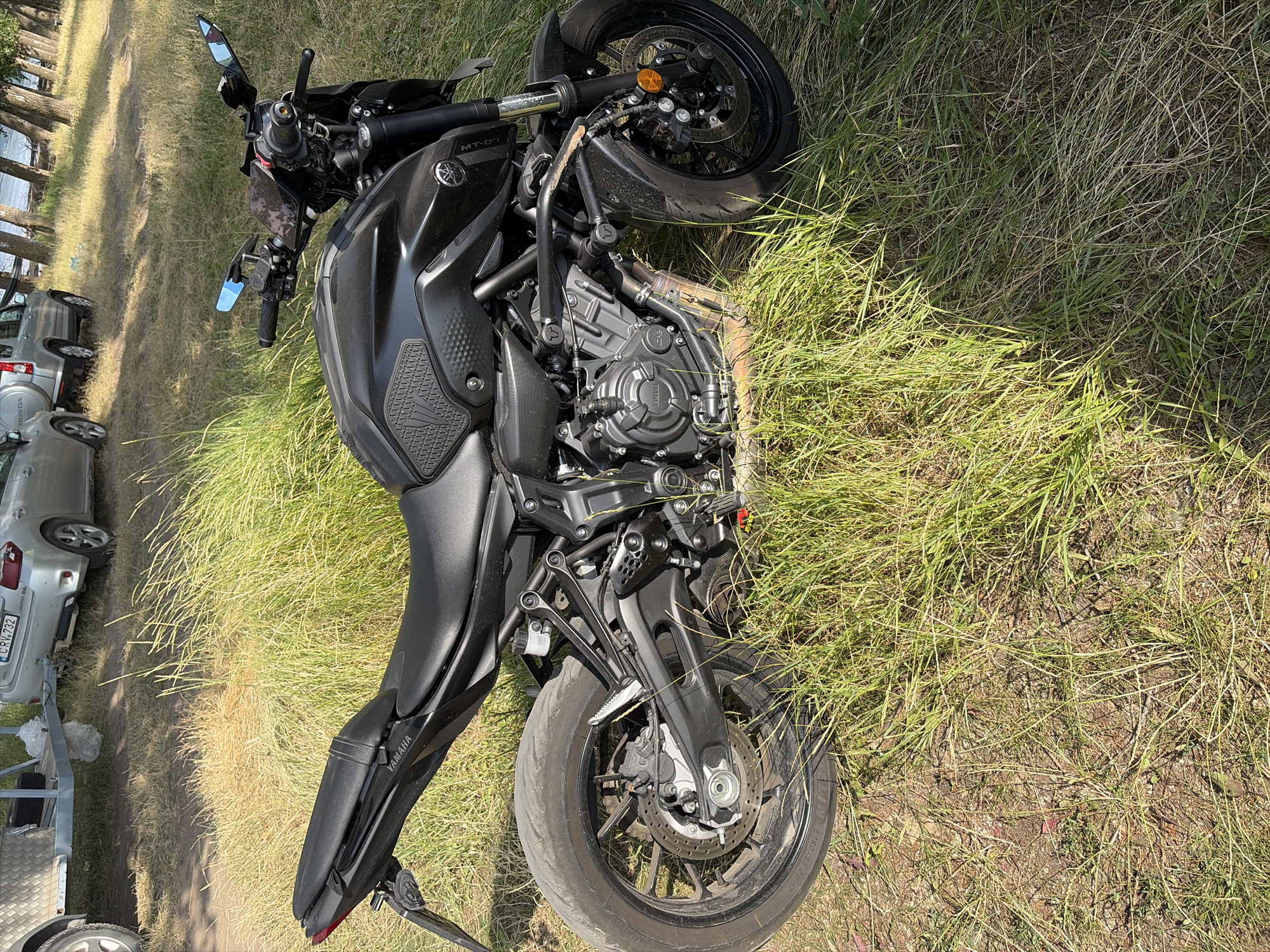 A black Yamaha motorcycle is parked on a grassy area. The motorcycle is facing to the left and its rear tire is visible. In the background, a silver car with the license plate CRV 732 is parked. Another silver vehicle is visible behind it. The scene appears to be in Oxentea, Moldova, near a body of water and a line of trees. The ground is covered with grass and dry leaves. The sun is shining, casting shadows on the ground.