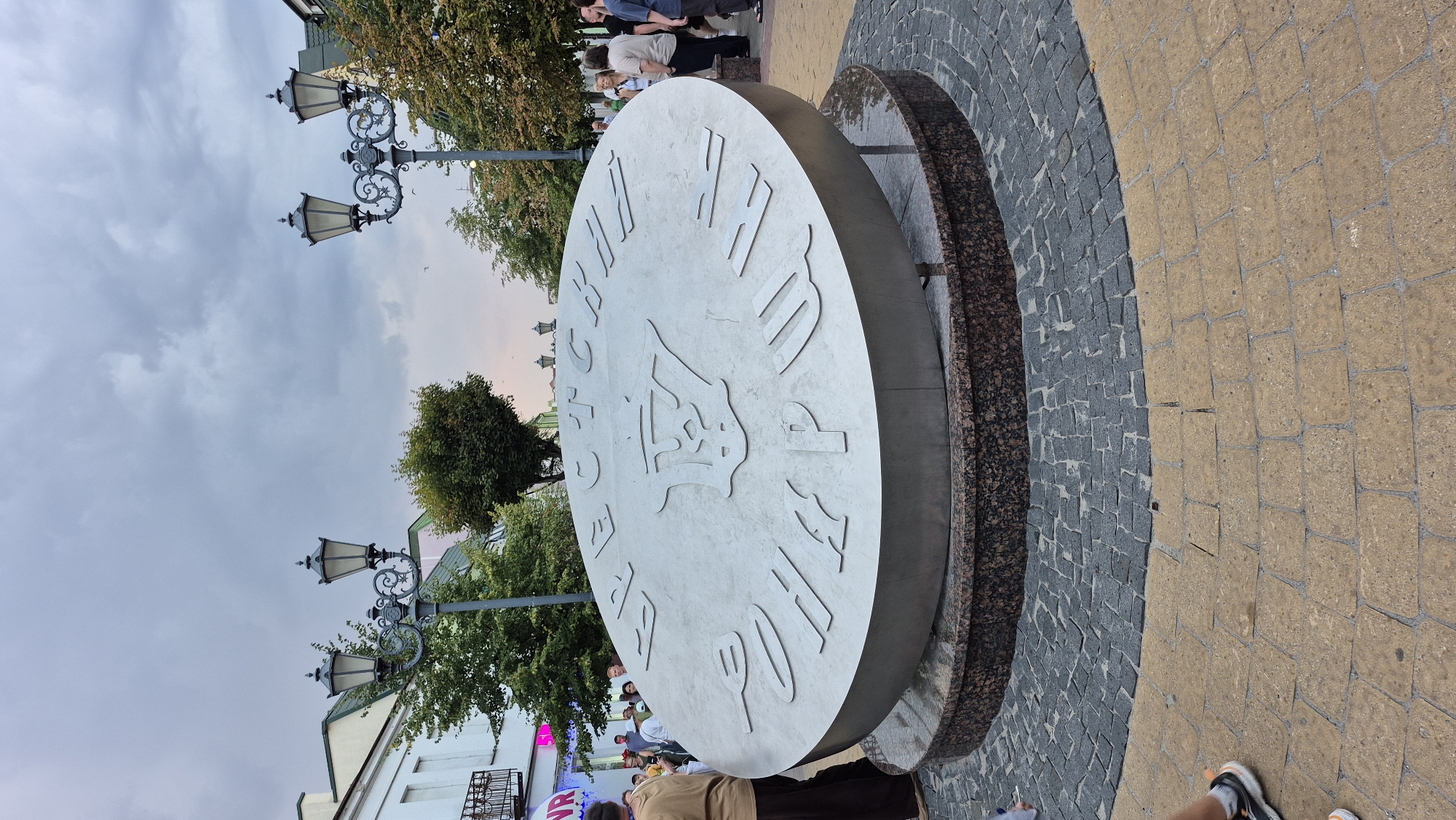In the city of Brest, Belarus, a large, circular monument stands in an outdoor plaza. The monument is made of a silver-colored material and features engraved Cyrillic lettering around its perimeter, as well as an emblem in the center. The monument is set on a tiered base, with the lower tier appearing to be made of dark, speckled stone and the upper tier of a darker, possibly granite material. The plaza itself is paved with a combination of dark gray and tan cobblestones arranged in concentric circles around the monument. Several people are present in the background, some observing the monument and others walking through the plaza. The environment appears to be an urban streetscape, with buildings and trees visible behind the monument. Ornate streetlights with multiple lamps are spaced along the street. The sky is overcast and cloudy.