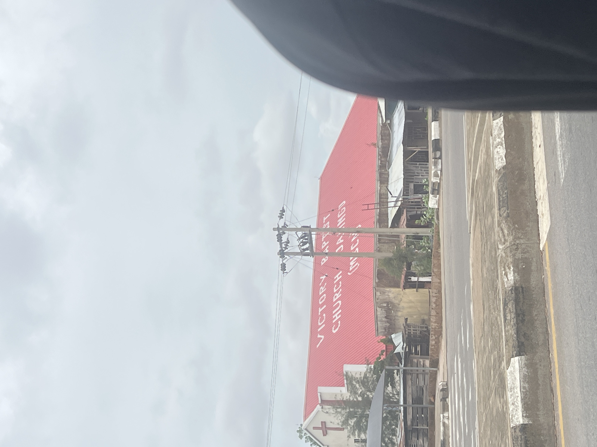 The image shows a view of Victory Baptist Church, Jalingo, in Nigeria. The church has a large red roof with white lettering that reads "VICTORY BAPTIST CHURCH JALINGO". A red cross is visible on the white facade of the building to the left. In front of the church, there are some small structures with corrugated metal roofs. A crosswalk is painted on the road in the foreground. The sky is overcast. The image is taken from inside a vehicle, as indicated by the dark blurred object on the right side of the frame.