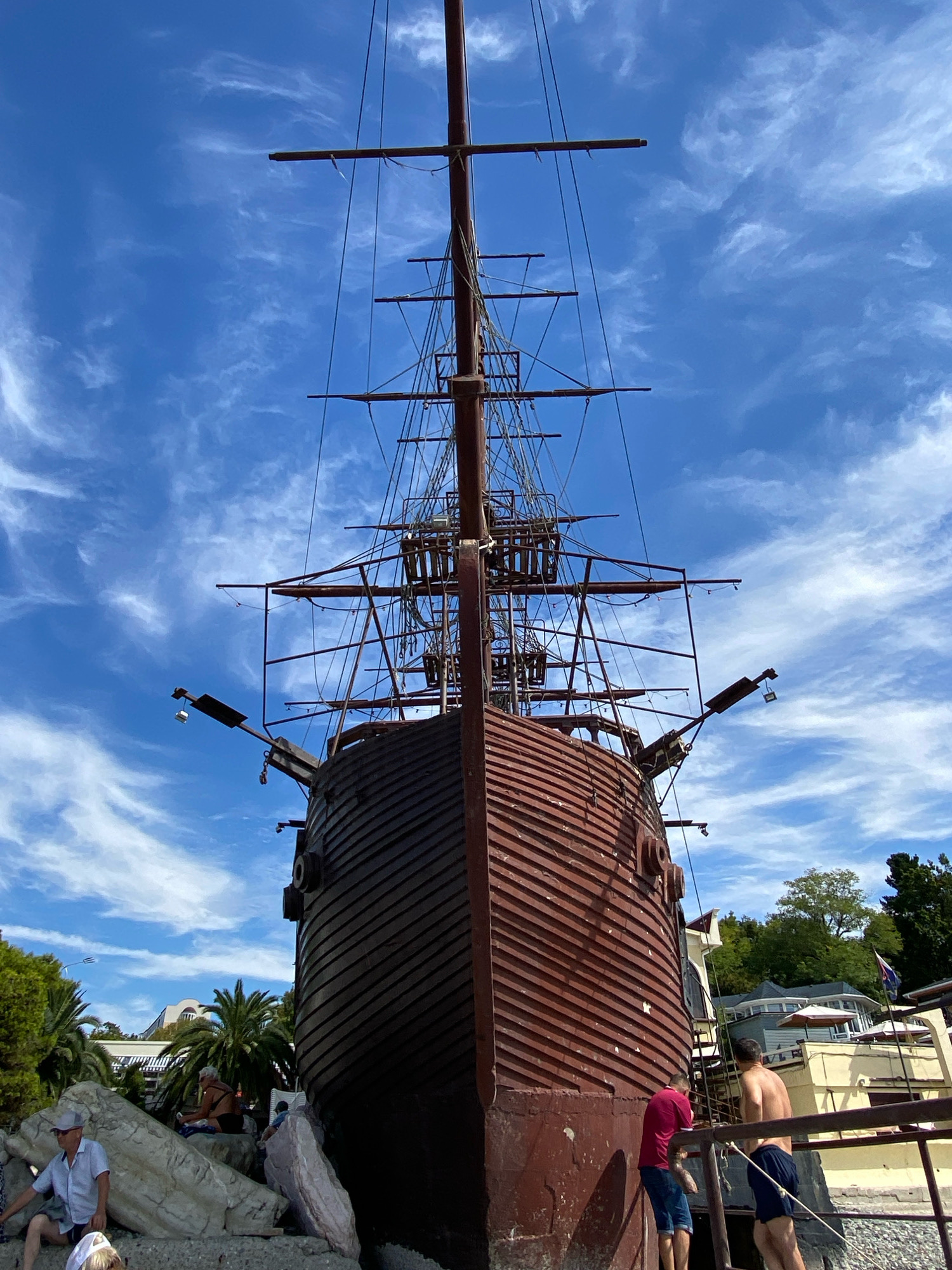 The image shows a view of a replica of an old sailing ship, likely a Galleon, positioned on land near the Black Sea coast. The ship's bow faces the viewer, and its tall masts and rigging are prominent against a bright blue sky with scattered white clouds. The ship's hull is dark red and appears to be constructed from wooden planks.
To the left of the ship, a group of people are visible near large rocks. One man wearing a light blue shirt and a grey hat is seated on the rocks, while another person's head is visible in the foreground. Further back, a woman is sitting on a blue towel. Palm trees and lush green foliage are visible behind the rocks, suggesting a warm climate.
To the right of the ship, a yellow building with white awnings and umbrellas can be seen. Two men are standing on a walkway in front of the building, with one wearing a red shirt and blue shorts, and the other shirtless wearing blue shorts. The walkway appears to lead to the ship.
The overall setting suggests a tourist attraction or a themed area, possibly in a coastal town or resort. Given the context provided, this scene is located in the city of Sochi, Russia, a popular Black Sea resort known for its subtropical climate and numerous attractions. The ship might be a permanent installation for entertainment or a historical exhibit.