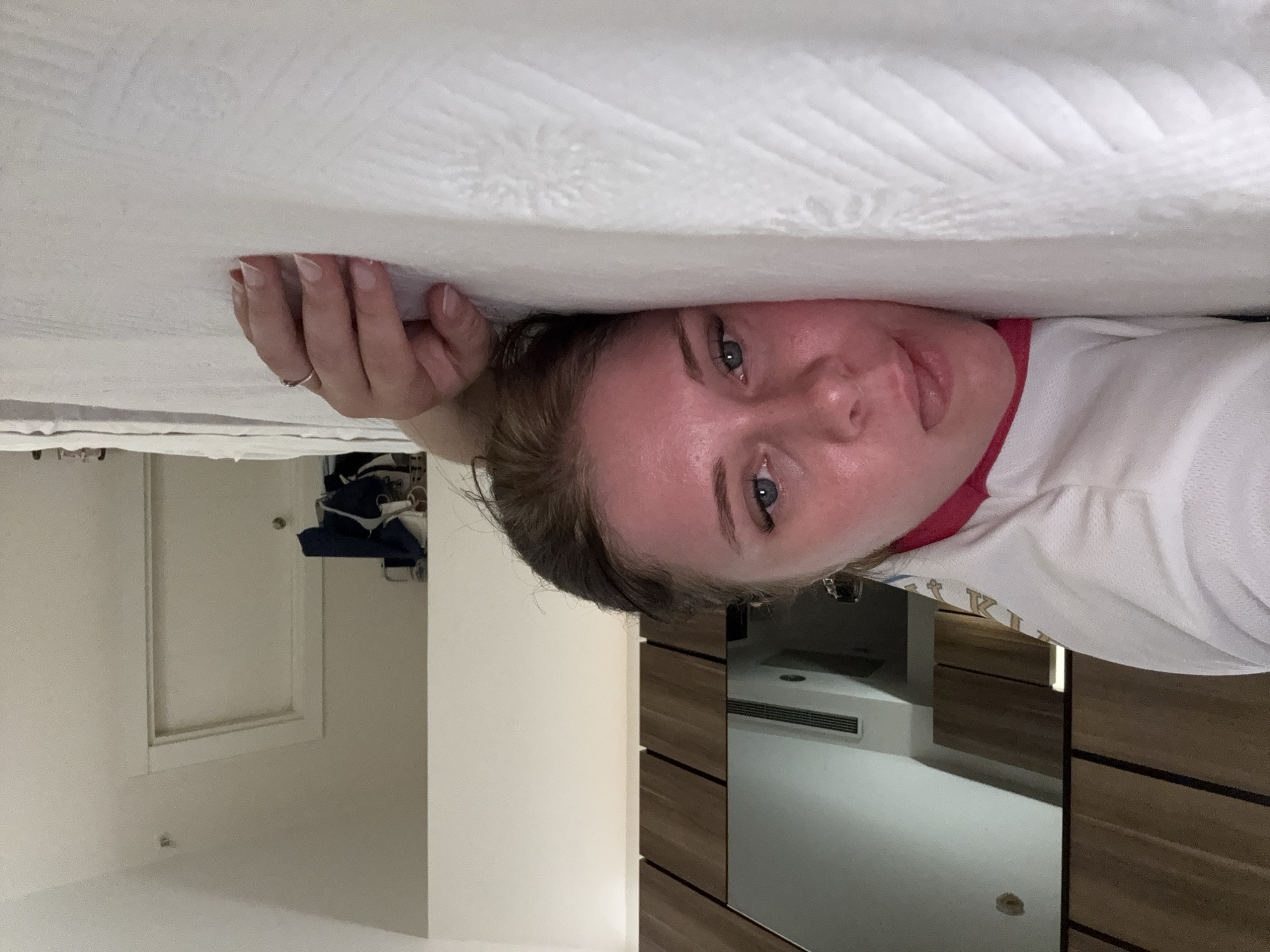 The image shows a person lying on a bed in what appears to be a hotel room. The person is facing the camera, with their head resting on the pillow and their body mostly out of frame. They are wearing a white top with a pink neckline. Their face is flushed, and their eyes are open, looking towards the camera. A hand with a ring is visible resting on the bedspread. The background shows a wooden wardrobe with a mirror, a door, and some clothing items. The bed is covered with a white, textured bedsheet. The overall lighting suggests an indoor setting, likely during the day. The location is identified as Aksu, Turkey, which suggests this is a hotel room in that city.
