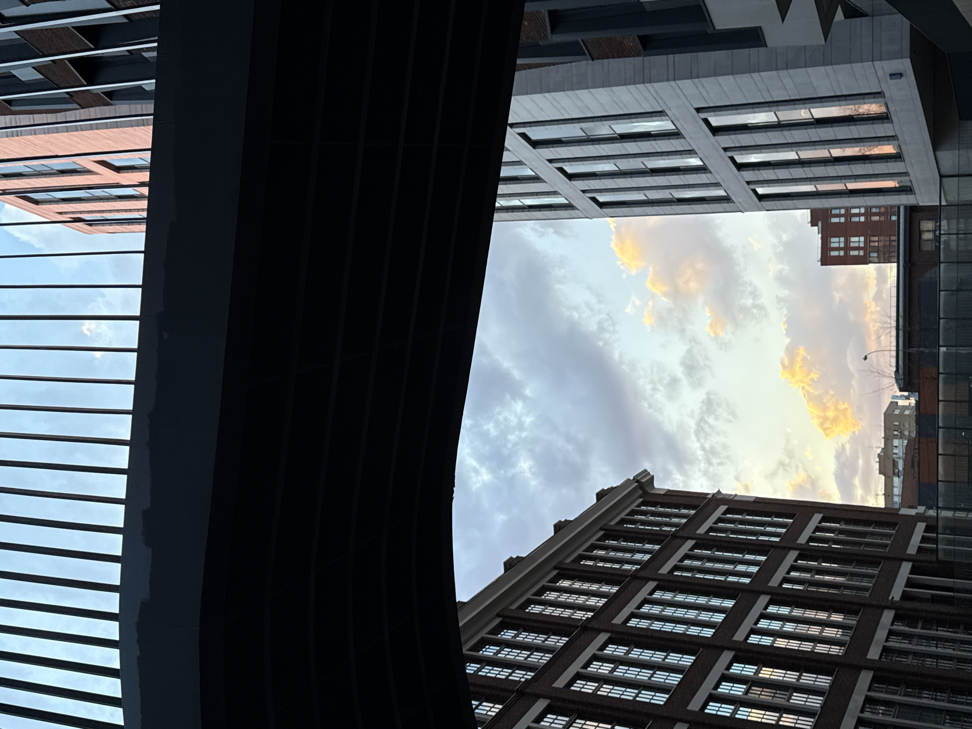 The image is a low-angle view of skyscrapers and sky. The buildings frame a patch of sky with golden clouds. The buildings are modern structures with many windows. One building has a brick facade, and another is grey concrete. A large, dark structure, possibly a bridge or elevated walkway, cuts across the left side of the frame. The perspective is looking upwards, emphasizing the height and scale of the urban landscape. It is not possible to confirm this is Moscow, Russia based only on the image.