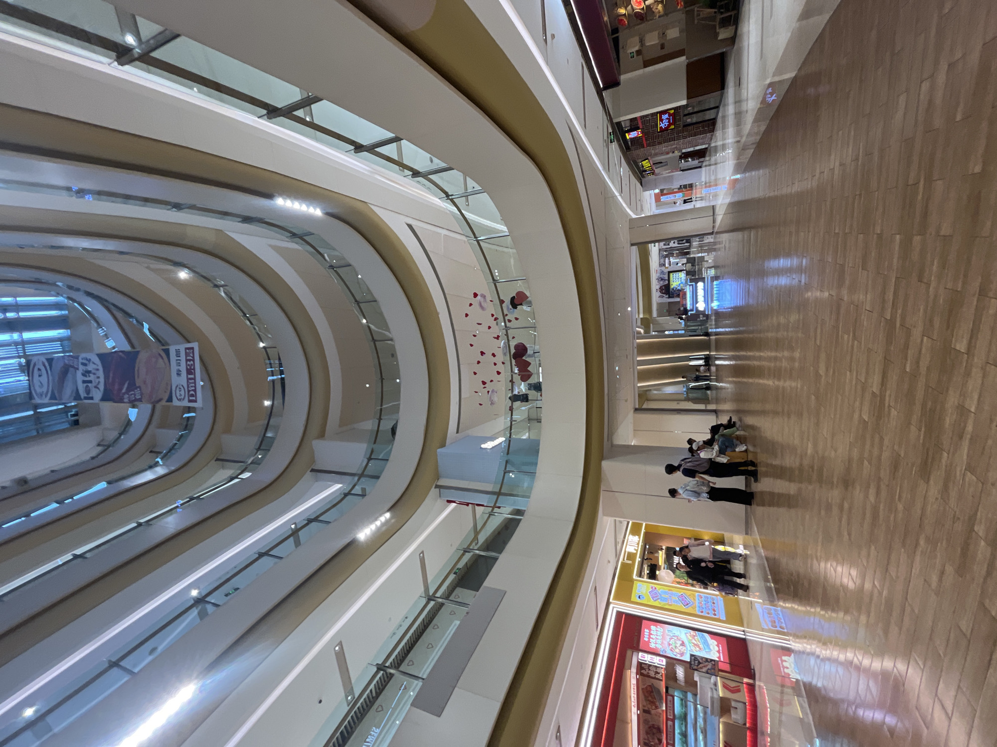The image depicts the interior of a multi-level shopping mall in Yuzhong District, China, with a wide-angle perspective looking upwards. The architecture features curved balconies on each level, creating an atrium-like effect. The interior is brightly lit by overhead lights, and sunlight streams in from above, reflecting off the polished wooden floor.
Several shops are visible on the lower levels, including one with a red facade displaying food items and another with yellow and blue signage. A group of people are gathered near the shops. Some are standing and looking at displays, while others are seated on a bench. A few individuals are walking through the mall's corridor.
The upper levels of the mall are lined with glass railings, and some decorative elements, such as red heart shapes, are visible on one of the balconies. The overall impression is of a modern and bustling commercial space.