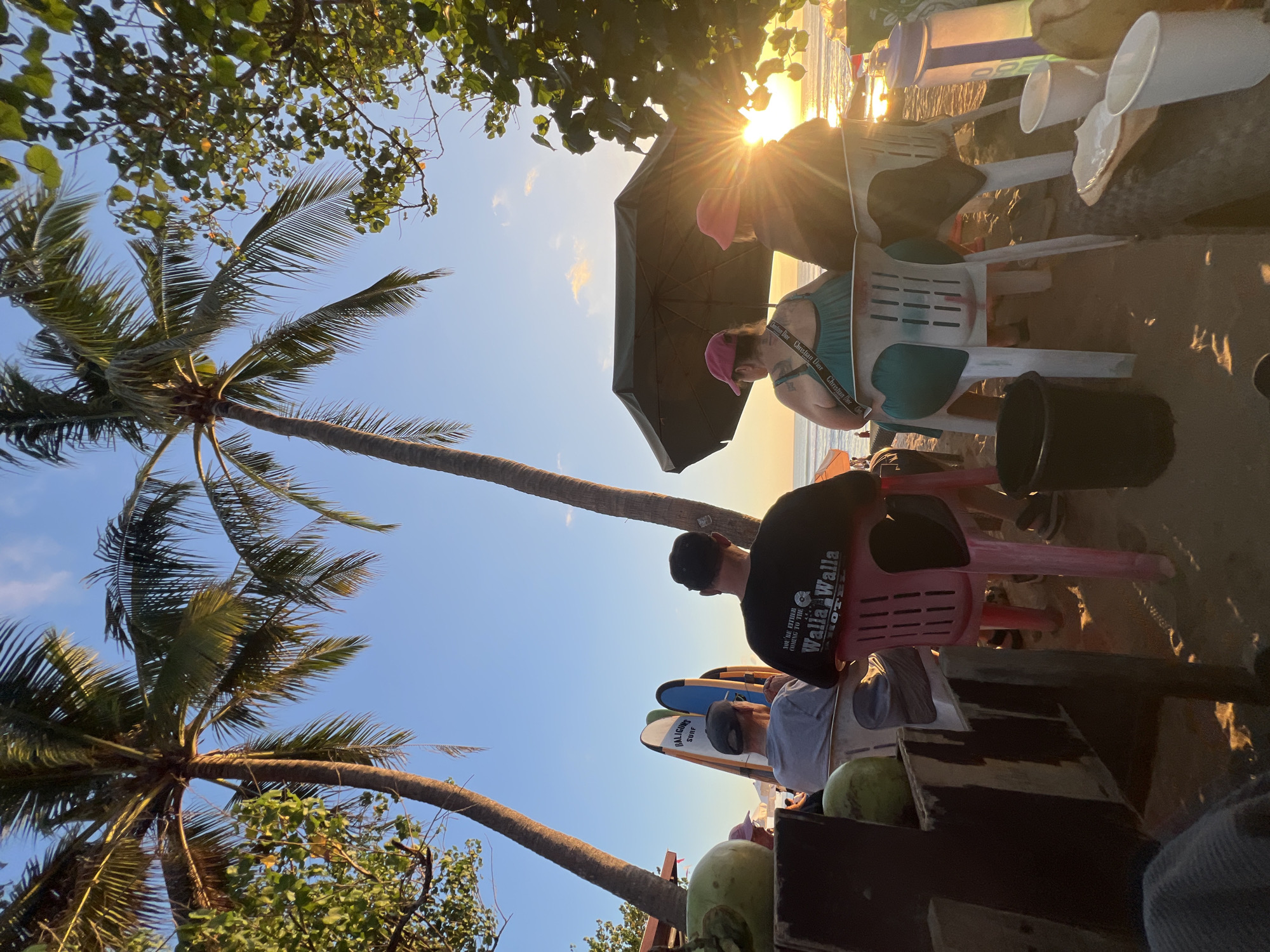 In Kuta, Indonesia, at sunset, four individuals are seated on plastic chairs on a sandy beach, facing a body of water. Two individuals are in the foreground, their backs to the camera, observing the ocean. One is wearing a black t-shirt with white text that reads "YOU'RE EITHER GOING TO THE WALLA WALLA HOTEL". The other is wearing a gray t-shirt and a baseball cap. Behind them, two people are partially visible, one in a teal dress and the other wearing a pink hat, both seated under a large umbrella.
Several surfboards stand upright behind the first two individuals, with "GALIGUNS SURF" written on one. To the left, a wooden structure holds two green coconuts. The environment is a beach with palm trees framing the scene against a clear blue sky with some scattered clouds. The sun is setting, casting a warm glow on the water and creating a lens flare effect. In the background, the ocean is visible with gentle waves. The sand is scattered with various objects, including plastic cups and a black bucket. The overall atmosphere suggests a relaxed evening by the sea.