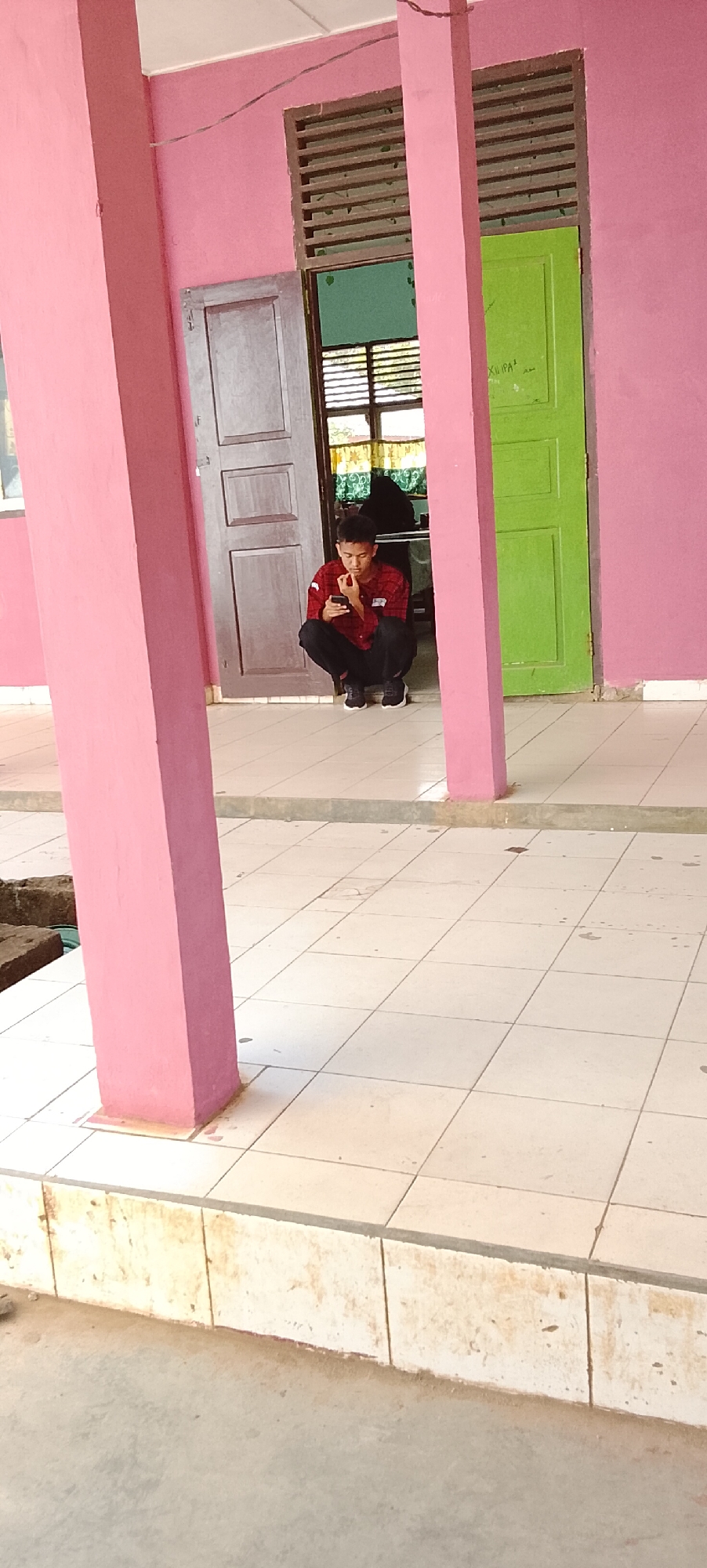 A young man is squatting on the tiled floor of a covered walkway, looking at his phone. He is wearing a red and black plaid shirt and black pants. The walkway has pink columns and a pink wall. Behind him are two doors, one brown and one green, with slatted windows above them. The floor is tiled with white squares. In the background, through the open doors, a classroom or office space is visible with desks and people. The scene appears to be in a school or institutional building, possibly located in Tanjung Setia, Indonesia.