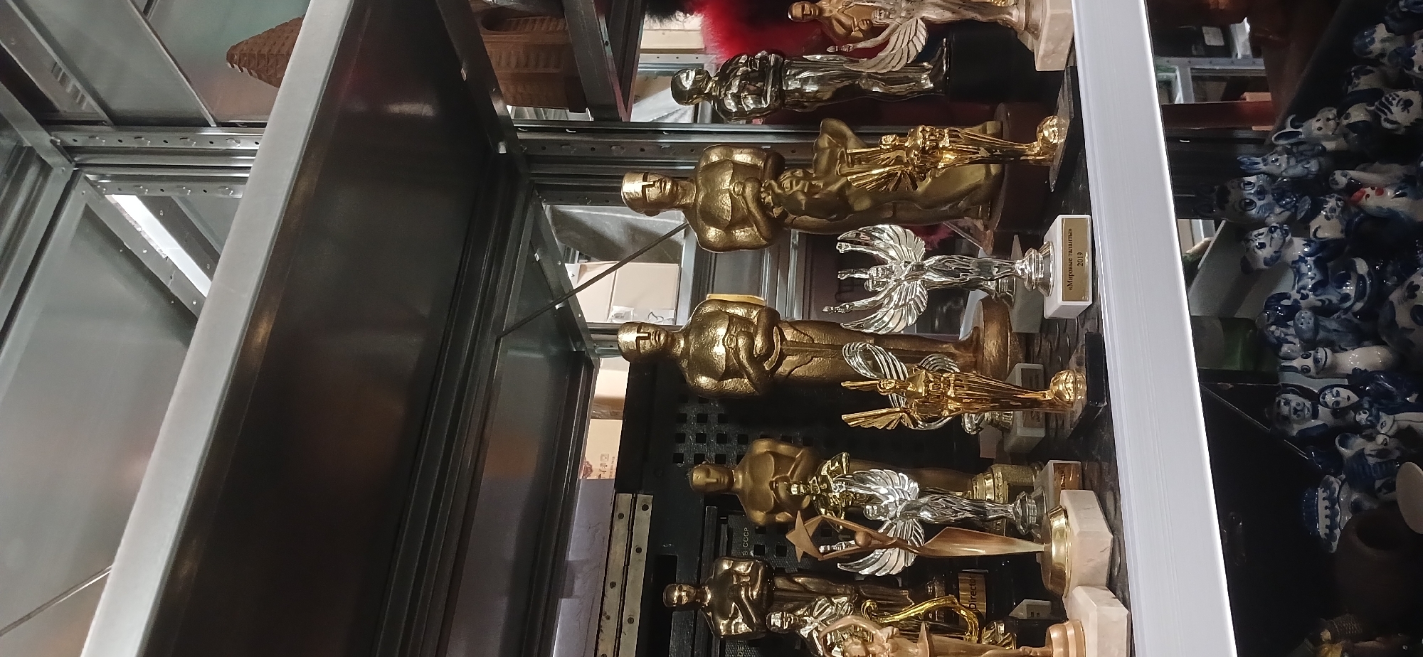 A high-angle, close-up shot displays a shelf inside a cabinet, presumably in Moscow, Russia, laden with statuettes and porcelain figurines. Several of the golden statuettes resemble Oscar awards, and some are paired with silver, winged figures on white bases. To the right of the statuettes are numerous small porcelain figurines predominantly in blue and white. The shelf appears to be part of a metallic storage unit, with sections of its structure visible on the left side of the frame.