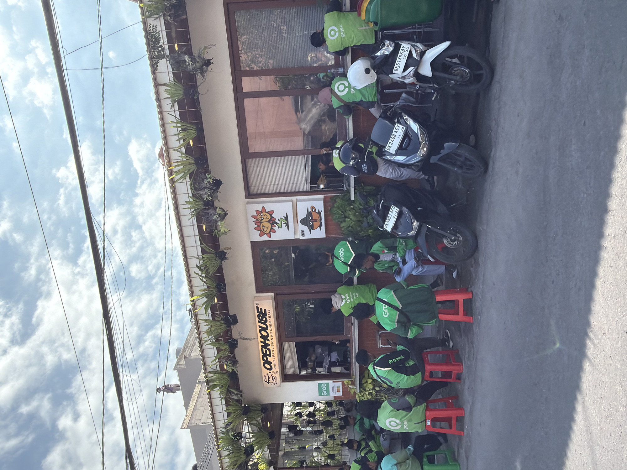 The image shows the storefront of "OPENHOUSE DAILY PROVISION KITCHEN BALI" in North Kuta, Indonesia. Several delivery drivers wearing green "Gojek" and "Grab" uniforms are seated on small red and green plastic stools outside the restaurant, presumably waiting for orders. Several motorcycles are parked nearby. The building's facade is decorated with plants. Utility wires are visible in the upper-left corner of the image. The sky is partly cloudy. License plates on the motorcycles indicate they are registered in Bali, such as "DK 5803 AFO," "DK2488 AEX," and "DK5139KAY." The time of year appears to be late in the year due to the license plate expiry dates.