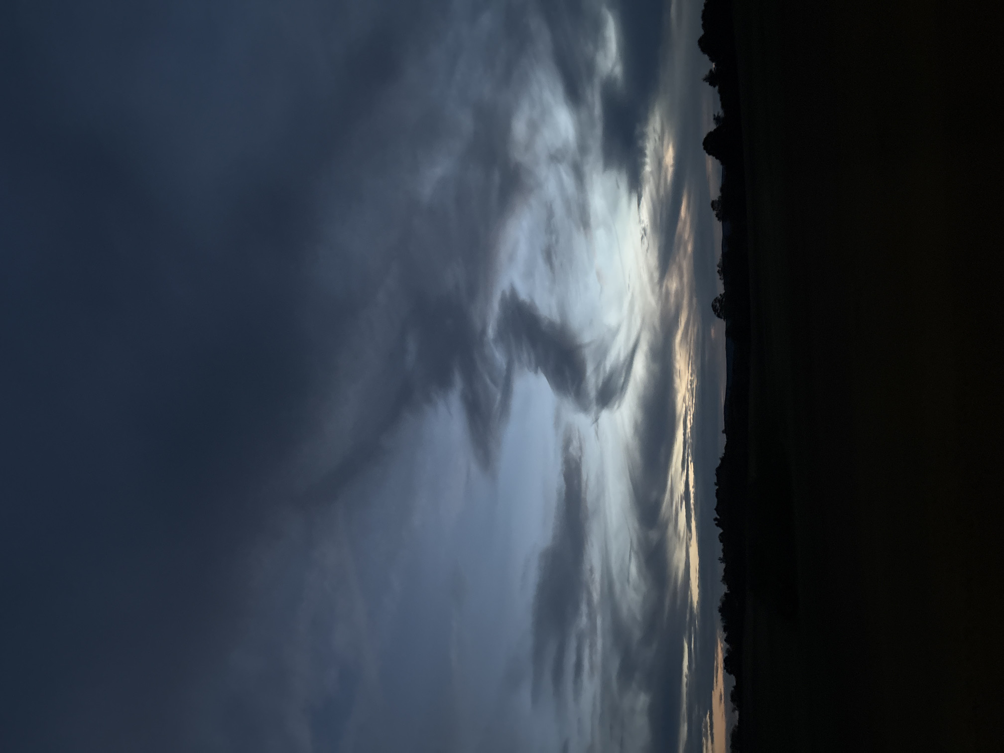 The image depicts a twilight scene, with the sky dominating the frame and a dark silhouetted landscape at the bottom. The sky is filled with dramatic, swirling clouds in shades of dark gray, deep blue, and lighter, ethereal blues and grays. There is a hint of warm, pale orange light on the horizon, suggesting the last moments of sunset or the pre-dawn twilight. The clouds are textured and layered, with some appearing wispy and others more dense and opaque.
The lower portion of the image is a dark, indistinct silhouette of a natural landscape. A line of trees and possibly rolling hills forms the horizon. The foreground is entirely dark, suggesting a field or open ground at night or in very low light conditions. The overall mood of the image is somber, dramatic, and peaceful. There are no people, animals, or man-made structures clearly visible.
