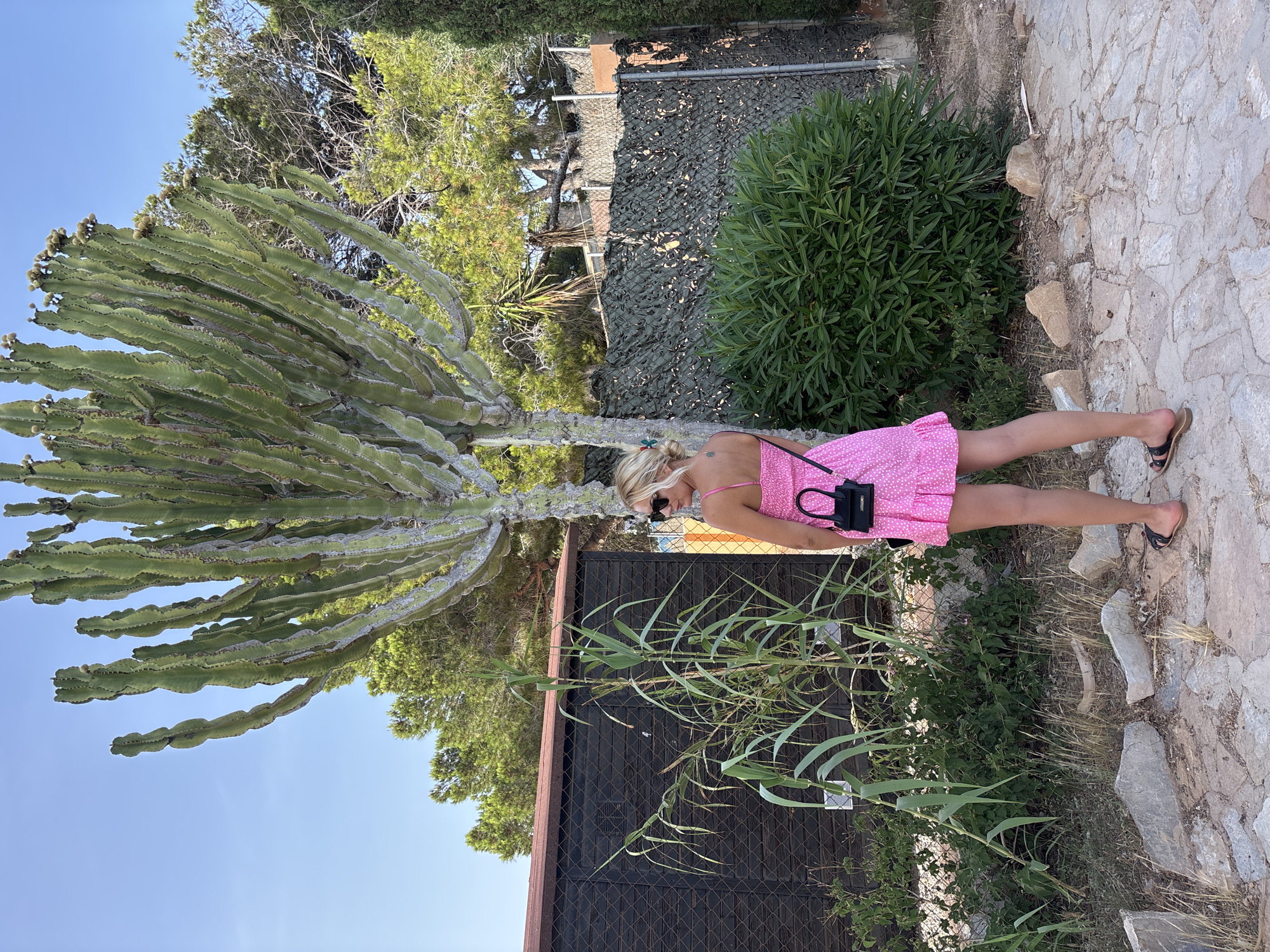 A blonde woman wearing a pink polka dot dress and black sunglasses stands in front of a large cactus. She is holding a small black purse. The background includes a dark wooden shed with a red roof, green foliage, and a chain-link fence covered with camouflage netting. The ground is covered with rocks and dry grass. The scene appears to be outdoors in a sunny climate. This location is in Orihuela, Spain.