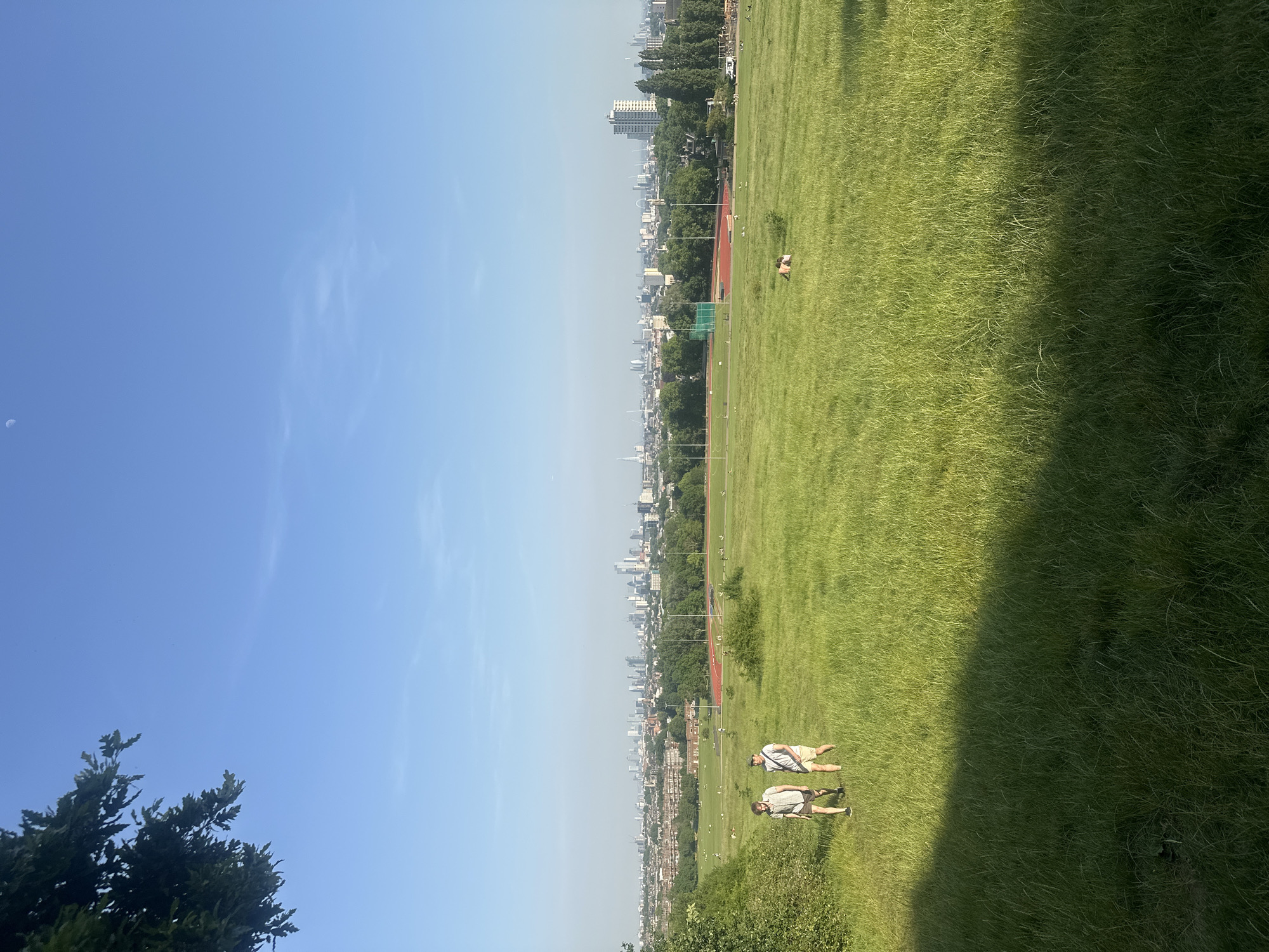 The image shows a green field in London, United Kingdom, with the city skyline in the distance. Two people are lying on the grass in the foreground, while another person is lying further away. A running track is visible near the city. The sky is blue with some clouds. Trees and bushes surround the field.