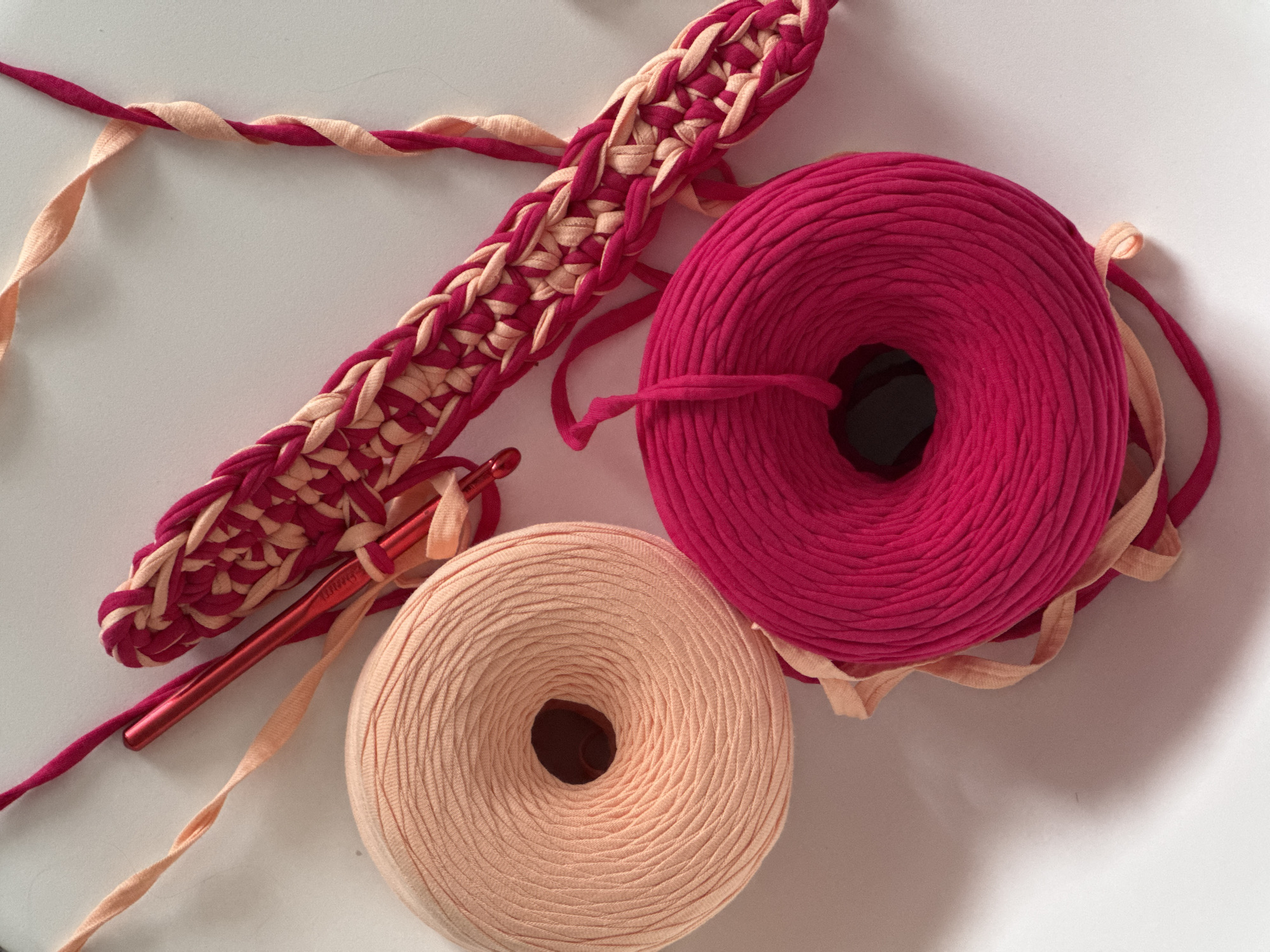 The image captures a crafting scene set on a clean, light-colored surface, likely indoors in Izhevsk, Russia. The primary subjects are two large spools of t-shirt yarn, one in a vibrant magenta or fuchsia hue, and the other in a soft, light peach or beige.
Adjacent to the yarn spools lies a partially completed, broad crocheted piece, showcasing an alternating pattern of both magenta and peach yarn, forming a decorative braid-like texture. A metallic red crochet hook is inserted into the stitches of this work, indicating an active or recently paused project. The brand name "PRYM" is visible on the handle of the crochet hook.
Loose strands of both yarn colors are visible, with some peach yarn casually draped over the magenta spool and a magenta strand extending from its center. The lighting is soft and diffused, suggesting a daytime setting without direct sunlight, casting subtle shadows that add depth to the composition. The overall impression is one of creative work in progress.