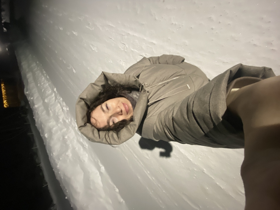 The image captures a nighttime scene in the city of Узяр, Russia, characterized by extensive snow cover. A single adult female is depicted in a close-up selfie, occupying the central and upper-mid frame. She has dark, curly hair and is wearing a light brown or taupe hooded winter coat, with the hood up. A dark scarf or neck garment is visible beneath the coat. Her eyes are partially closed, and her mouth is slightly upturned, indicating a smile. Her left arm is extended towards the camera, holding the device. A dark shadow of her and the extended arm is visible on the snow to her left. The ground is entirely covered in snow, displaying both smooth and disturbed textures. Snowbanks line what appears to be a cleared path or road. In the background, against the dark night sky, artificial lights are discernible, including a horizontal line of warm, yellow lights on the far left, possibly decorative, and blurred white lights further down the path, likely streetlights. The environment is consistent with a winter evening.