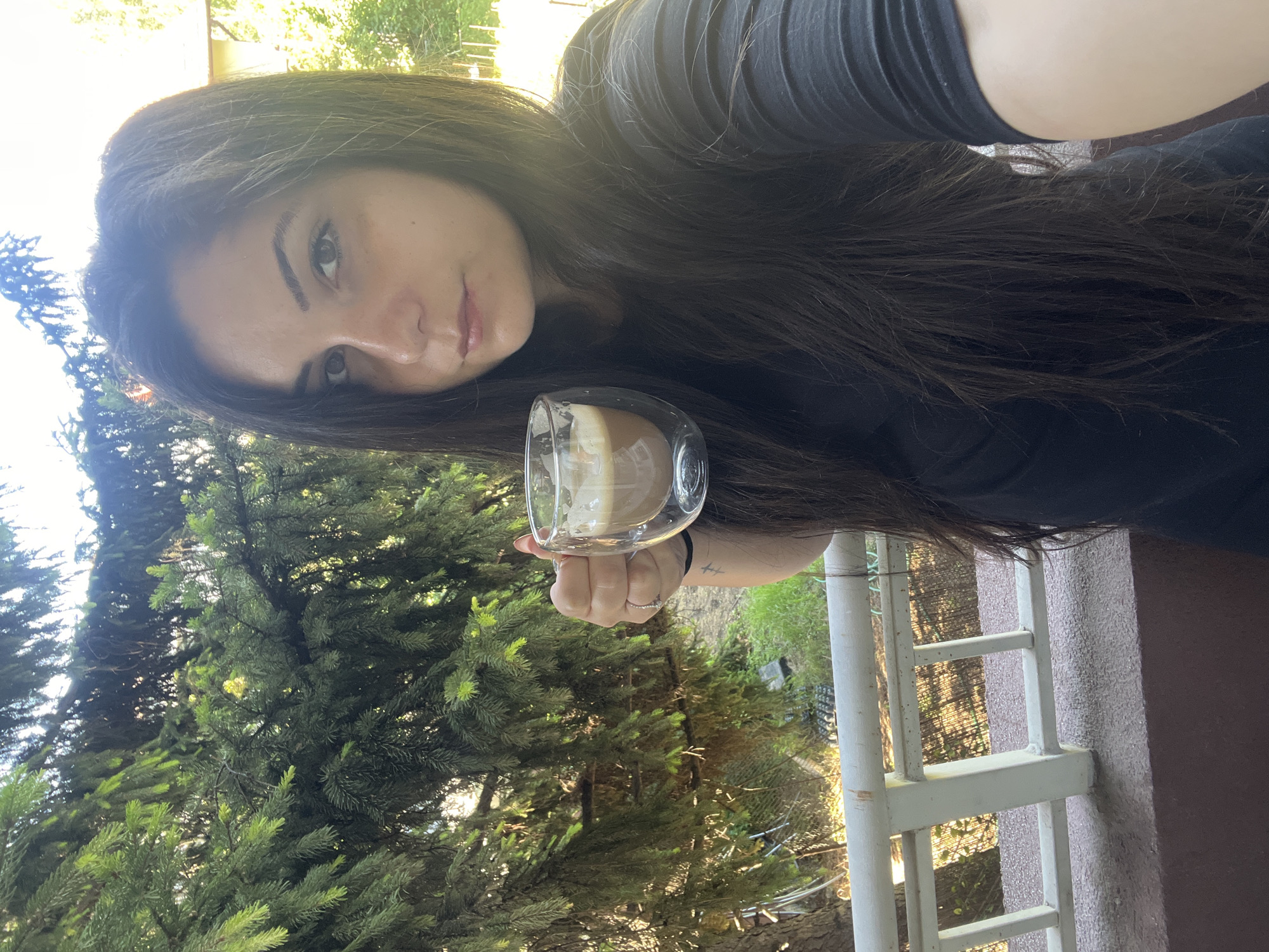 A woman with long, dark hair stands on a balcony, holding a clear mug filled with a brown beverage topped with foam. She is wearing a black t-shirt. Behind her, lush green pine trees frame the scene, with sunlight filtering through the branches. In the background, a building is visible. The balcony has a white railing. The overall impression is a relaxed, outdoor moment. The location is the city of Sofia, Bulgaria.