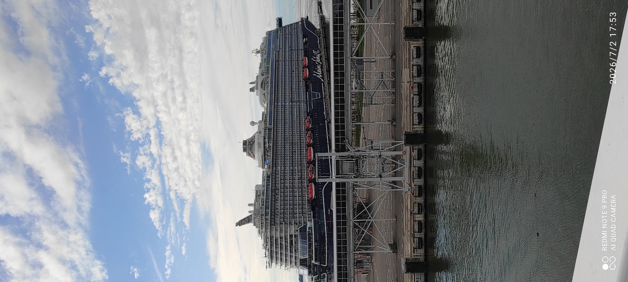 This image depicts a large, dark blue cruise ship docked at a pier in Tallinn, Estonia. The ship's name, "Mein Schiff," is visible on its side. The vessel is multi-decked and appears to be at port, with people possibly on board or disembarking, though no individuals are clearly visible. Orange life rafts are mounted along the ship's hull.
The setting is a harbor with a calm, dark body of water on the right side of the frame, reflecting the sky and the ship. The pier itself is made of concrete and has industrial infrastructure, including metal gantries and bollards. The background shows a bright blue sky with scattered white clouds, suggesting fair weather. The lighting indicates it is daytime, possibly late afternoon due to the angle of the sun.
Visible text includes the ship's name, "Mein Schiff," and a timestamp in the upper right corner that reads "2026/7/2 17:53". Additionally, a watermark in the lower right corner reads "REDMI NOTE 9 PRO AI QUAD CAMERA". The overall impression is one of a typical port scene, with the cruise ship being the central focus. There are no discernible emotions conveyed in the image.