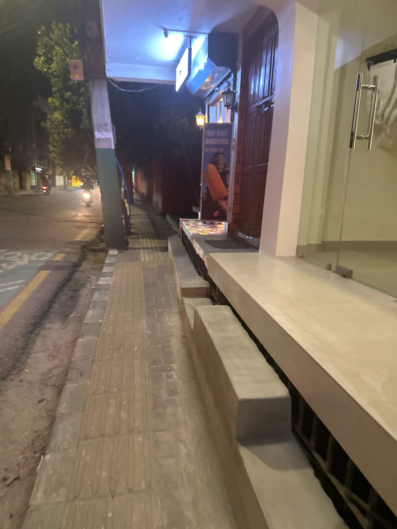 The image captures a street-level view at night in Lalitpur, Nepal, focusing on a sidewalk adjacent to a building. The sidewalk is constructed with textured concrete tiles, designed for tactile paving, and bordered by a road. A few motorcycles are visible on the road, with one rider in the foreground. The building features a wooden door and a glass facade, with a sign advertising "real fruit popsicles." Lighting from the building and streetlights creates a moderately illuminated scene. The environment appears urban, with the presence of a sidewalk, road, and commercial establishments.