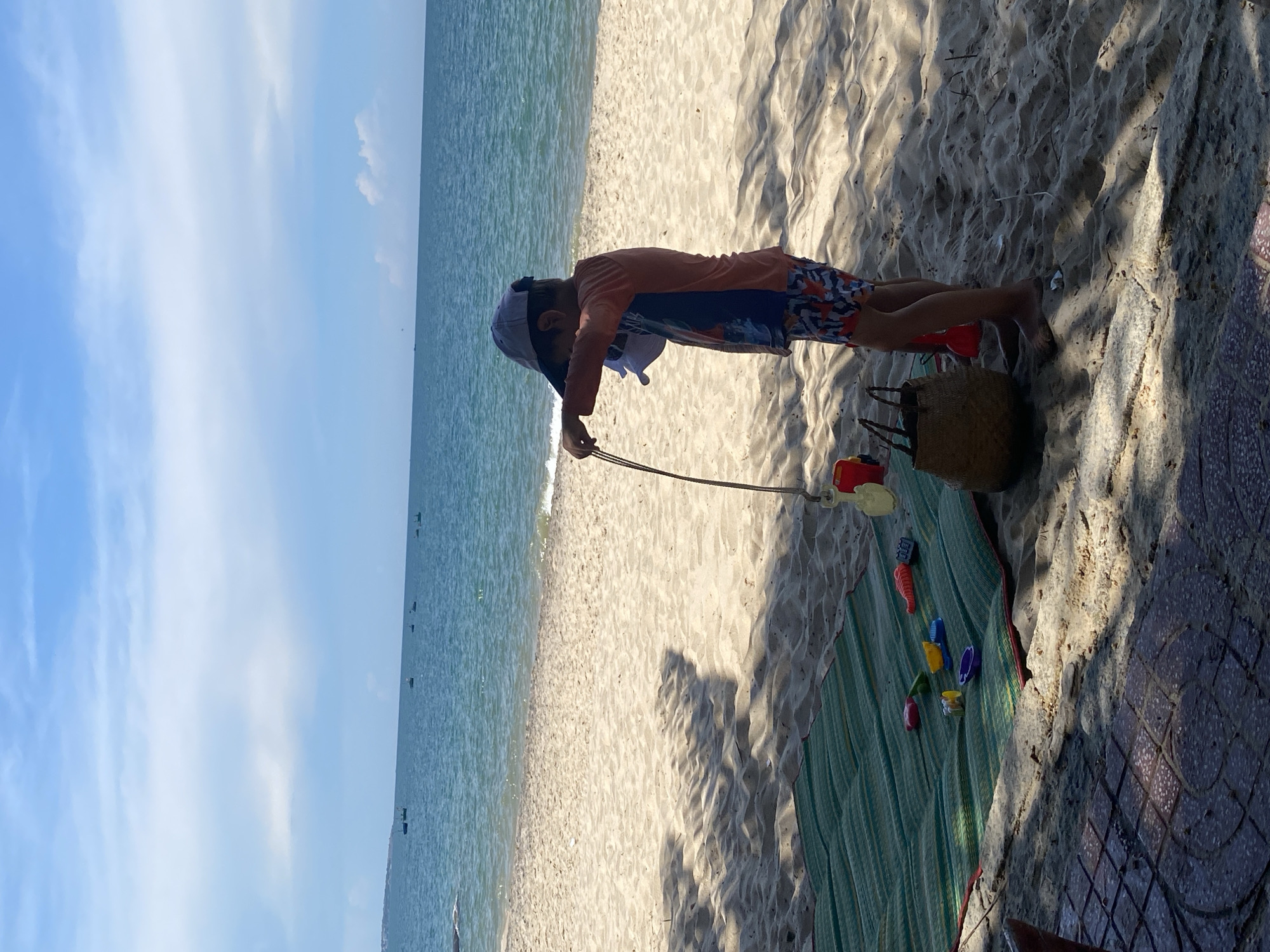 The image depicts a sun-drenched beach scene, likely in Phu Thuy Ward, Vietnam, under a clear blue sky with scattered clouds. A young child, wearing a light-colored cap, an orange rash guard with blue accents, and patterned swim trunks, stands on the sandy shore. The child is holding a thin, dark string attached to a pale yellow toy bucket and possibly a shovel, looking down as if playing or examining the toy.
In front of the child, a green woven mat is spread on the sand. Scattered on the mat are various colorful plastic beach toys, including red, green, yellow, purple, and blue items, along with what appears to be a red bucket and spade set. A straw beach bag rests on the sand near the mat. The ocean is visible in the background, a calm expanse of blue water with small waves gently lapping the shore. Several small fishing boats are dotted on the horizon. The foreground shows a paved area with a patterned surface, partially in shadow cast by unseen trees or structures. The overall atmosphere is one of leisure and a typical day at the beach.