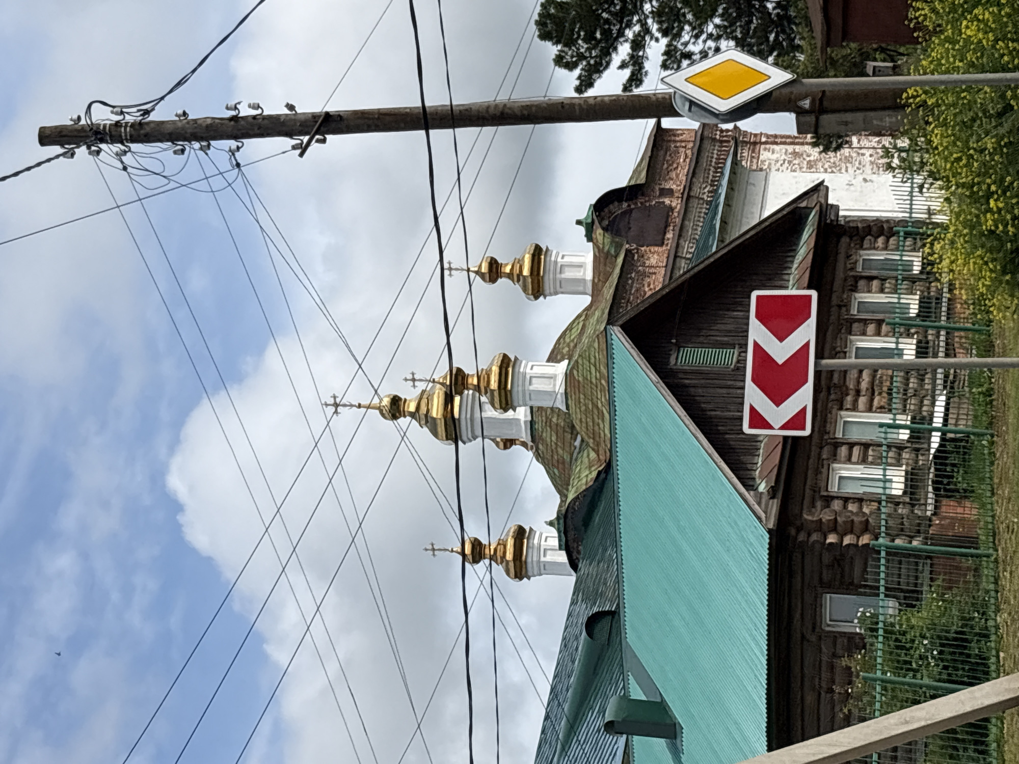 The image depicts a street scene in the city of Kstynino, Russia, featuring a wooden house with a turquoise metal roof in the foreground. To the left of the house, a tall, weathered wooden utility pole is visible, supporting numerous electrical wires that crisscross the sky. A traffic sign with a red chevron pointing left is mounted on a pole in front of the house. Behind the wooden house, the gilded domes and crosses of a Russian Orthodox church, characterized by white and brick architecture, rise against a cloudy sky. The background includes trees and more utility poles. The ground in front of the house is covered with green grass and flowering yellow plants, with a green metal fence separating the property from the street. A small bird is visible in the upper left portion of the sky.