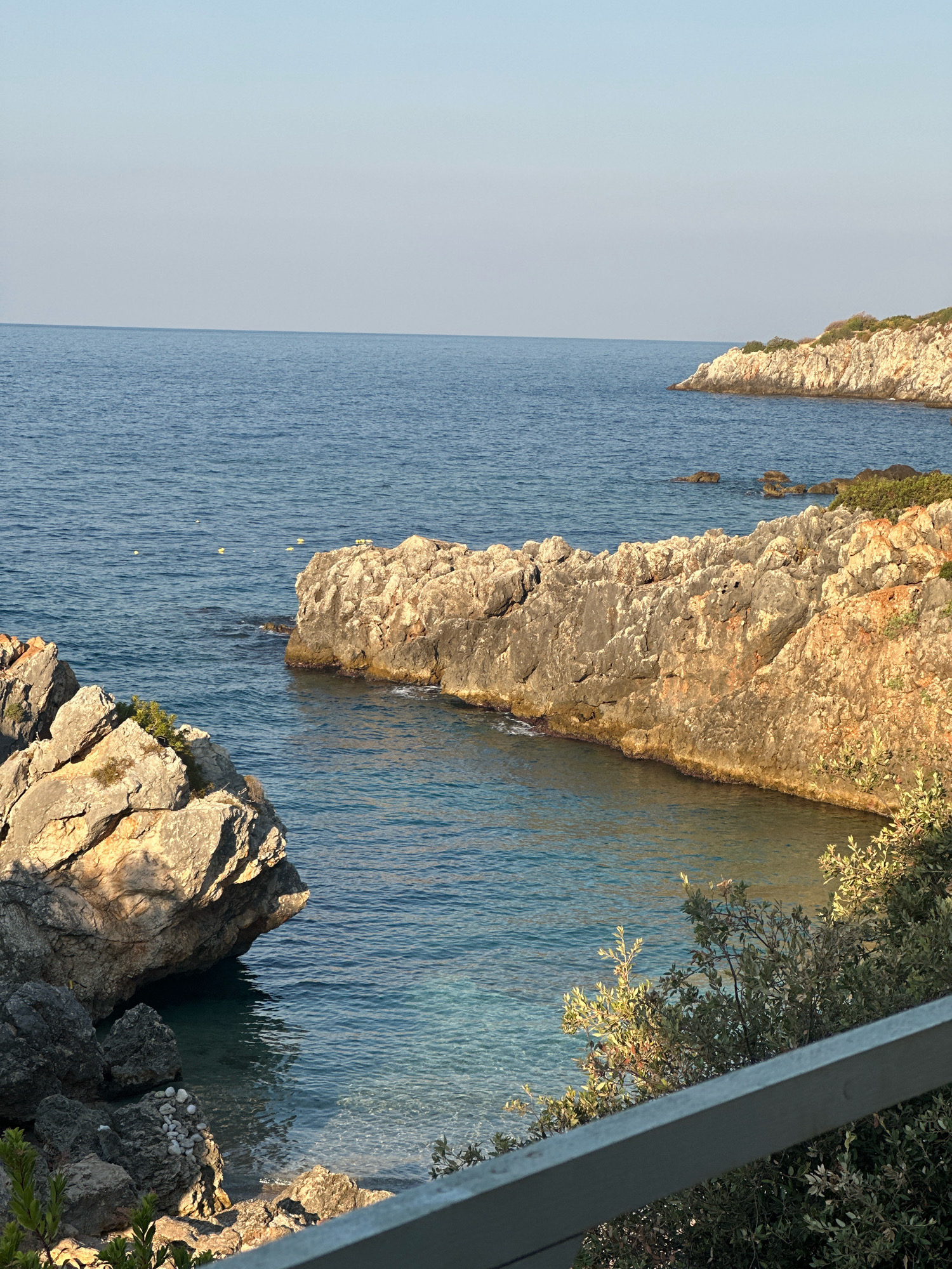 An eye-level shot captures a coastal view with the sea and rocky formations. The calm, blue water fills the majority of the frame, with a clear horizon line where it meets a pale blue sky. To the right, a rugged, rocky coastline juts out into the sea, adorned with sparse green vegetation. In the foreground, more rocky outcrops are visible, with the clear, shallow water revealing the seabed near the shore. A grey railing runs along the bottom right of the image, suggesting a vantage point from a balcony or walkway. The scene is bathed in natural light, indicating it's daytime, likely morning or late afternoon due to the warm tones on the rocks. There are no people or animals visible in the frame.