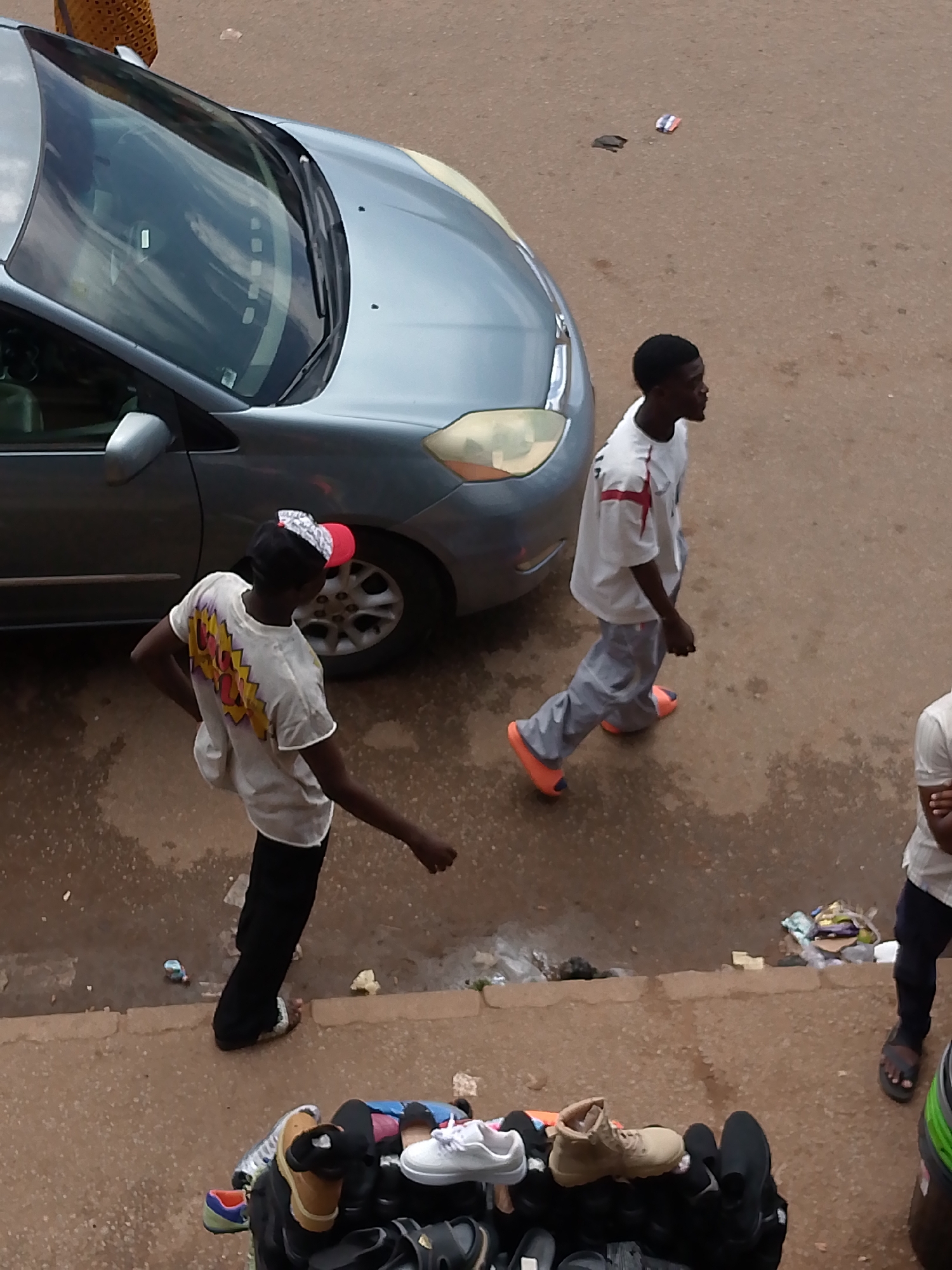 In Bukuru, Nigeria, two young men are walking on a dusty, reddish-brown street next to a silver minivan. One man, on the left, wears a white t-shirt with a colorful graphic on the back, black pants, and a cap with a red and white brim. He is captured mid-stride, with his left arm bent at the elbow and his right arm extended slightly backward. The second man, on the right, is walking in the same direction, slightly ahead and to the right of the first. He is wearing a white t-shirt with red accents and light gray pants, and bright orange slip-on shoes. His arms are down by his sides. Further to the right, the lower body of a third person is visible, wearing dark pants and dark sandals. In the foreground, a collection of shoes is displayed on a surface, including boots, sneakers, and sandals. The environment appears to be an outdoor market or street setting, with scattered debris visible on the ground.