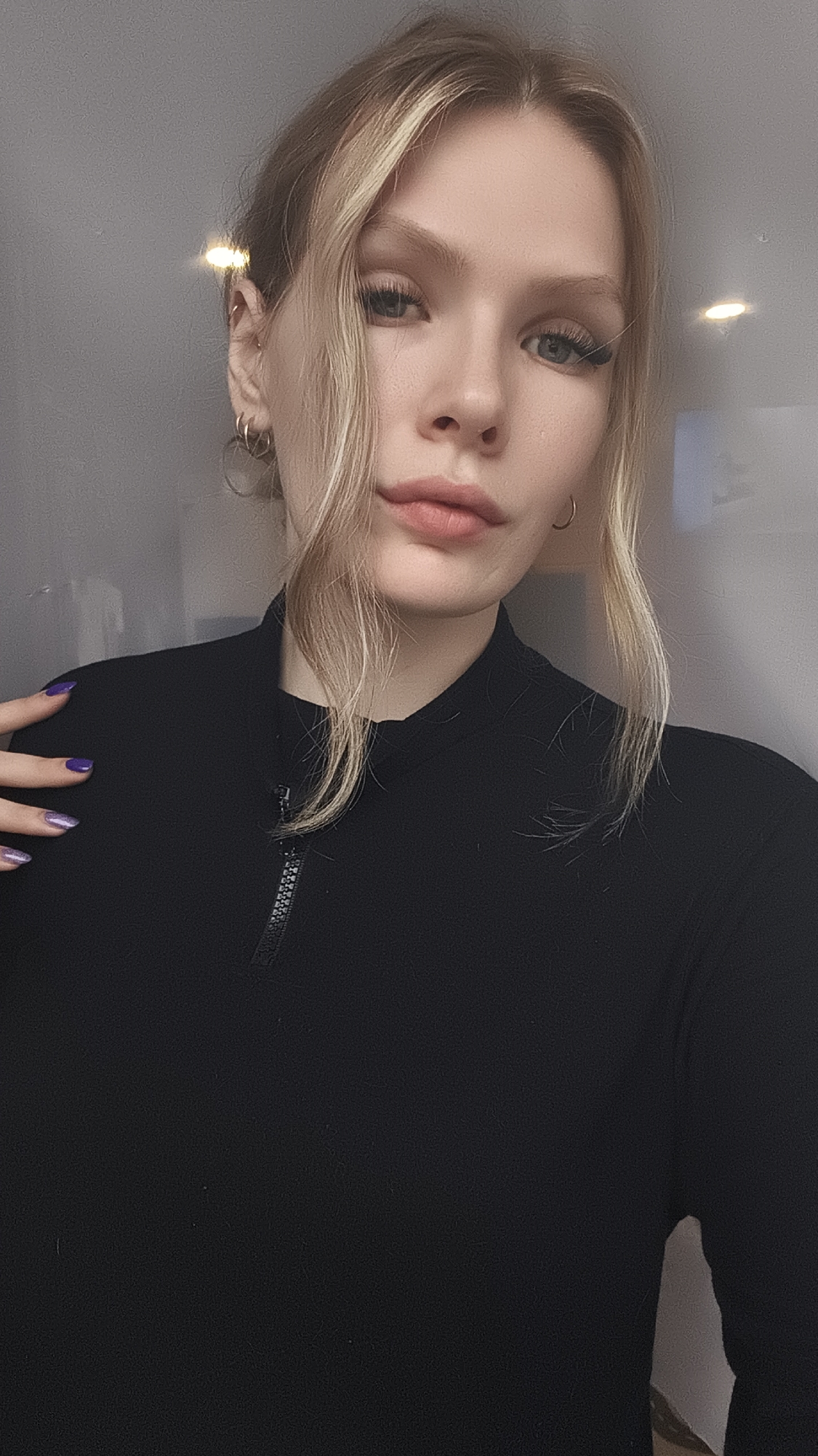 A woman with blonde hair and fair skin is posing indoors. She wears a black turtleneck shirt with a visible zipper. Her nails are painted purple. She is wearing multiple hoop earrings on her left ear. The background appears to be a plain wall with overhead lighting. The location in the city of Русский Вожой, Russia cannot be confirmed.