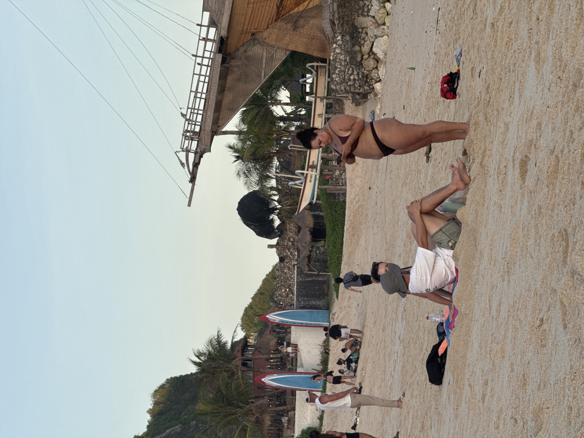 The image shows a beach scene in Kuta Selatan, Indonesia. People are on the sandy beach, some sunbathing and relaxing. There is a woman in a bikini using a smartphone. Behind her is a wooden structure, possibly part of a building or decoration. The beach is next to a building and behind some trees. Two surfboards are on the beach. The sky is overcast.