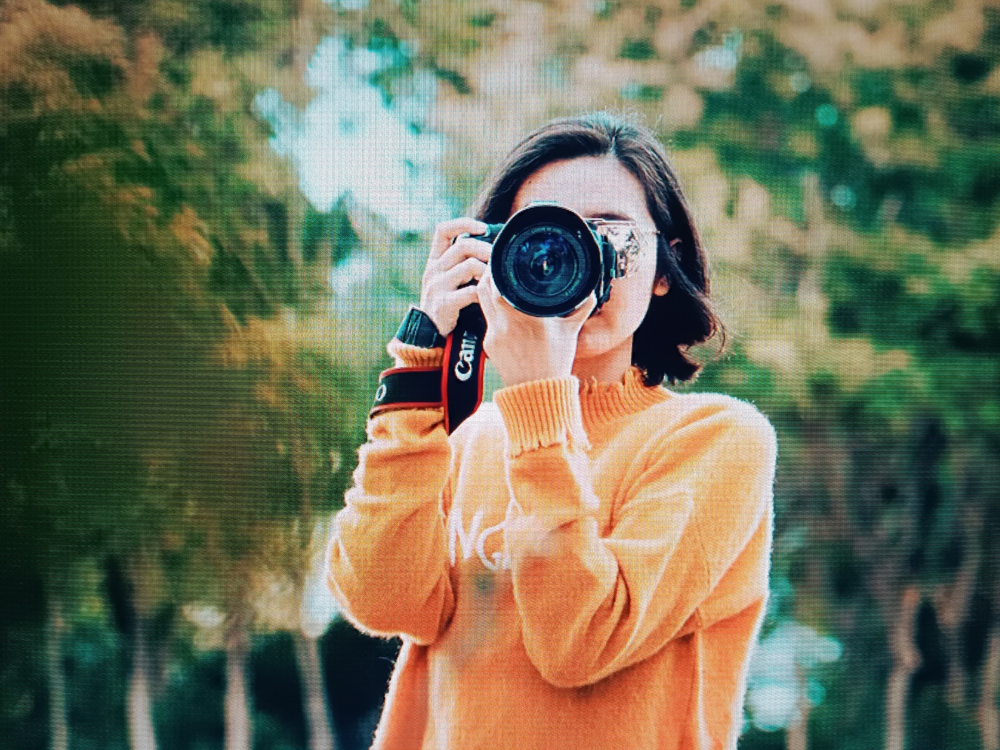 A young woman with short dark hair stands outdoors, holding a Canon camera to her face as if taking a picture. She is wearing sunglasses and a light orange sweater. The background is blurred, suggesting a natural setting with trees and foliage. The camera strap has the Canon logo visible. The location is not discernible from the image and there is no evidence indicating that it is Tbilisi, Georgia.