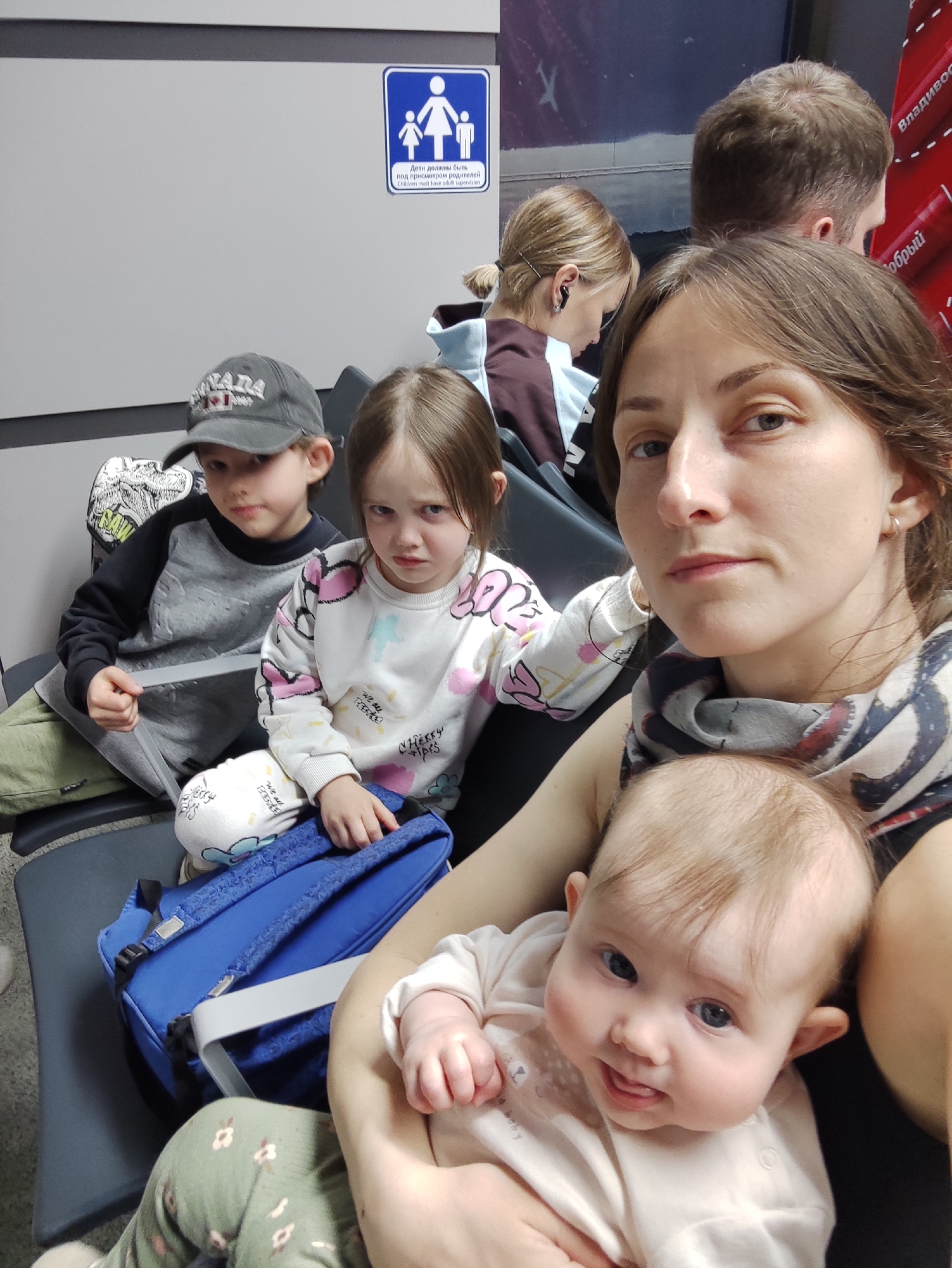 The image shows a woman holding a baby, sitting on a dark gray seat with three other children. A boy with a dark gray baseball cap sits to the left. A girl in a white sweater sits next to him and slightly behind him, appearing unhappy. A blue backpack sits near the girl. Behind them, a sign is visible that depicts a man, woman, and child holding hands. The text on the sign in both Russian and English states, "Children must have adult supervision". Further in the background, other adults sit or stand. A red advertisement is visible in the upper right corner. The woman and children are at a waiting area, likely an airport, at Sochi, Russia.