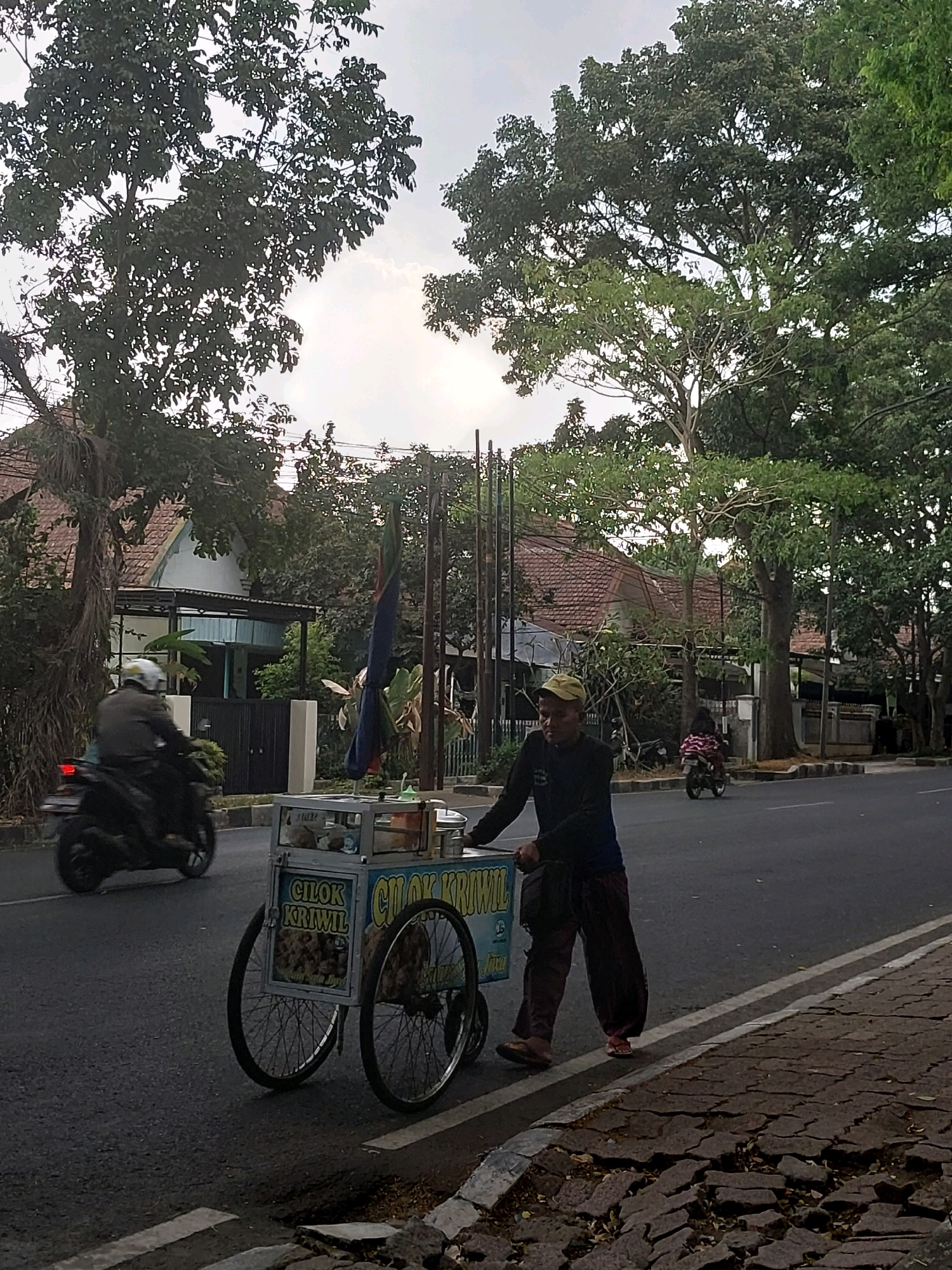 A street vendor is pushing a food cart on a road in Malang, Indonesia. The vendor, wearing a cap, long-sleeved shirt, and loose-fitting pants, is walking on the right side of the road, next to the curb. The food cart is a wheeled structure with a glass display case and signage that reads "CILOK KRAWIL". A motorcycle is passing by on the left side of the road. In the background, there are houses with tiled roofs and lush green trees lining the street. The sky is overcast, suggesting either early morning or late afternoon. The sidewalk in the foreground is made of cracked paving stones.