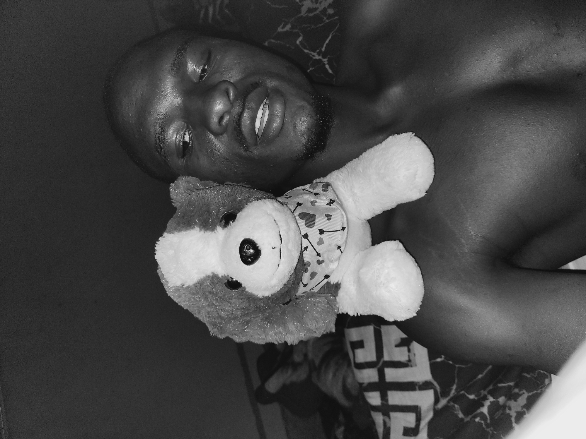 A black and white, close-up, high-angle selfie shows a dark-skinned person with a shaved head and a goatee lying down with a stuffed dog perched on their left shoulder. The person's mouth is slightly open, and their eyes are looking upwards and to the left. The stuffed dog is light-colored with a darker muzzle and ears, and it wears a bandana patterned with small hearts and arrows. The background consists of a dark, textured surface and a patterned blanket. The image appears to have been taken indoors, possibly in a bedroom, in Bukuru, Nigeria.
