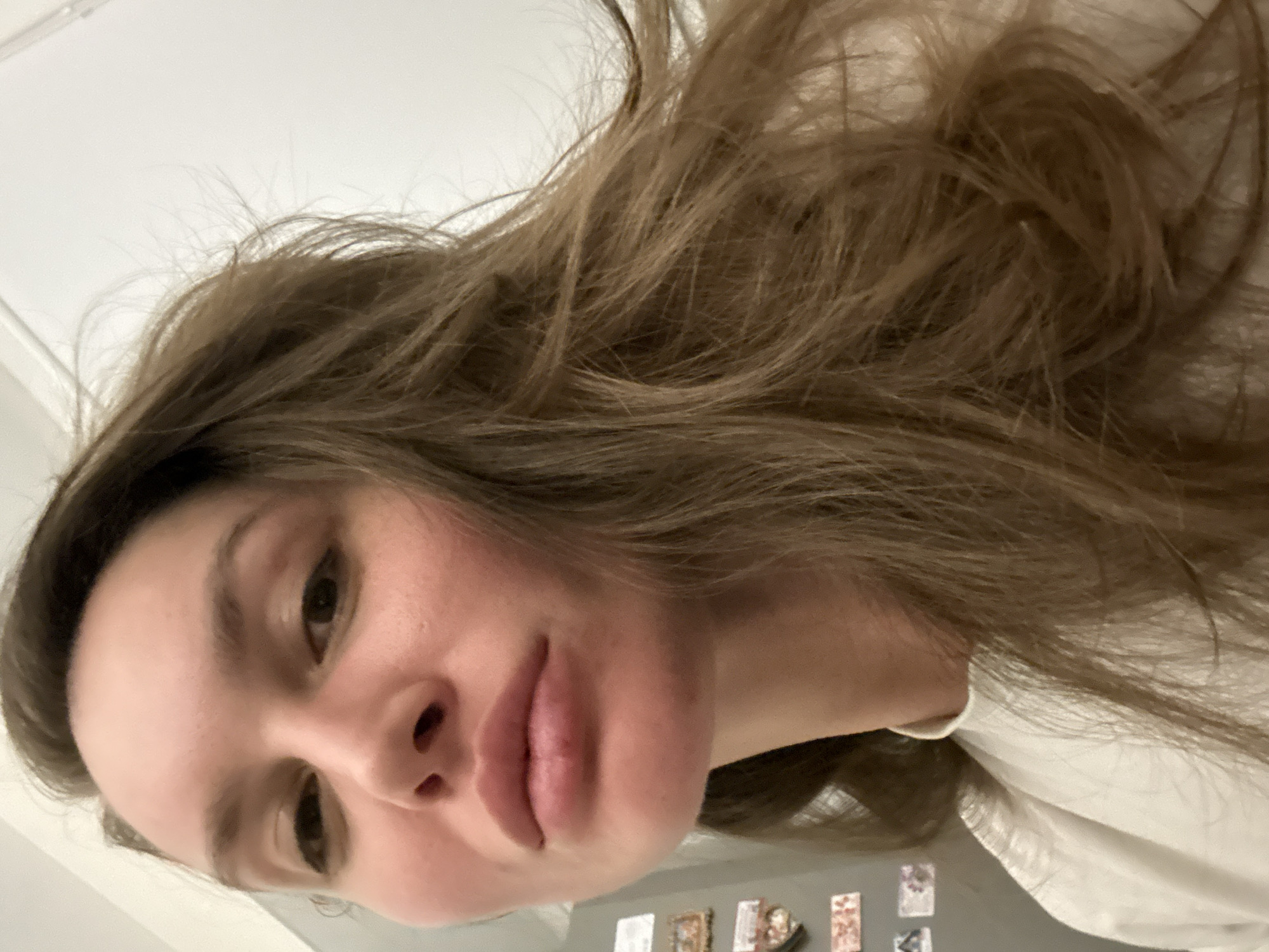 A selfie of a fair-skinned woman with long, light brown hair. She is wearing a white shirt. The background includes a white wall and a refrigerator decorated with magnets. The location is likely an indoor setting, but the city of Moscow, Russia cannot be determined from this image.